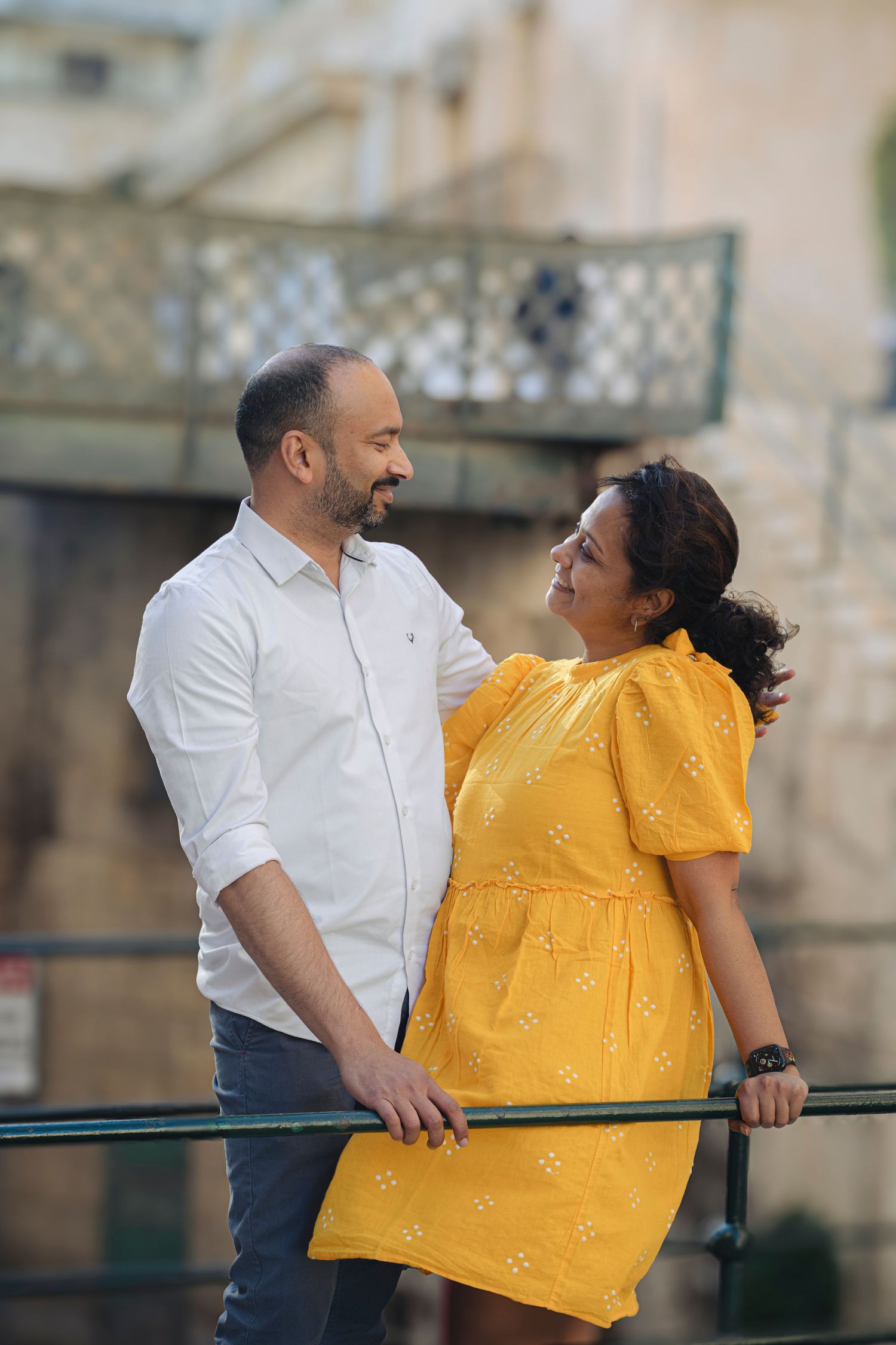 A couple in a romantic pose by a railing, woman in yellow dress, man in white shirt, smiling at each other outdoors.