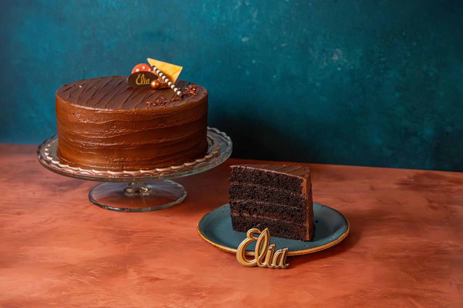 Chocolate layer cake slices on plates against a dark teal background with decorative gold accents.