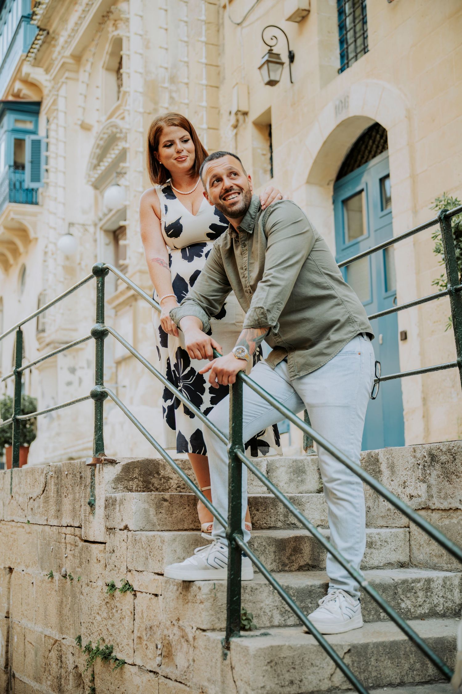 Couple posing on historic stone steps with ornate railings in a Mediterranean town.