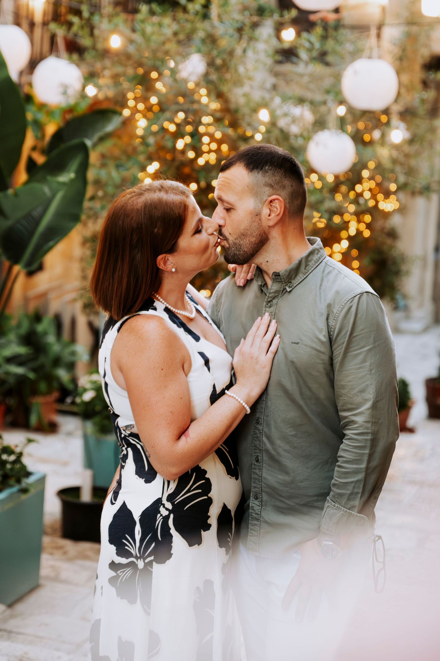 Romantic couple sharing a kiss surrounded by warm fairy lights and lush greenery.