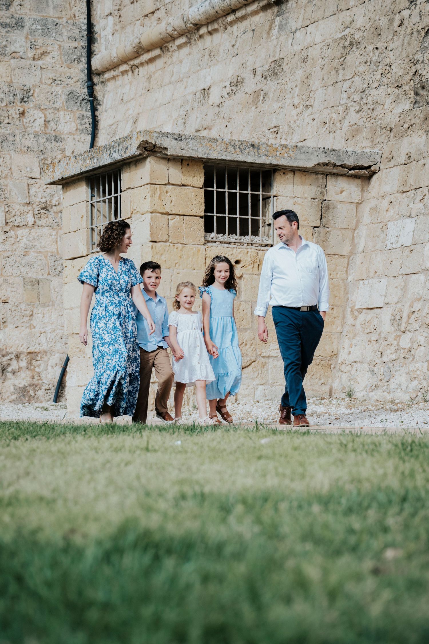 Family of five walking together outdoors near a stone wall, parents and three children dressed in blue and white.