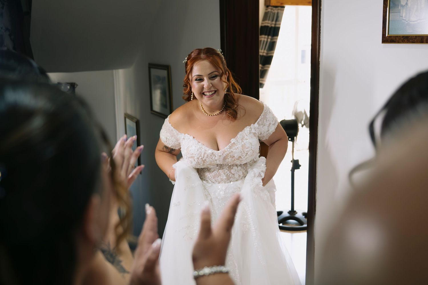 Bride in off-shoulder lace wedding gown smiling as guests applaud during bridal preparation.