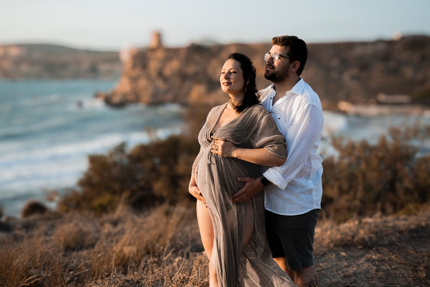 Pregnant couple embracing on coastal cliffside at golden hour, with ocean waves and historic ruins in the background.
