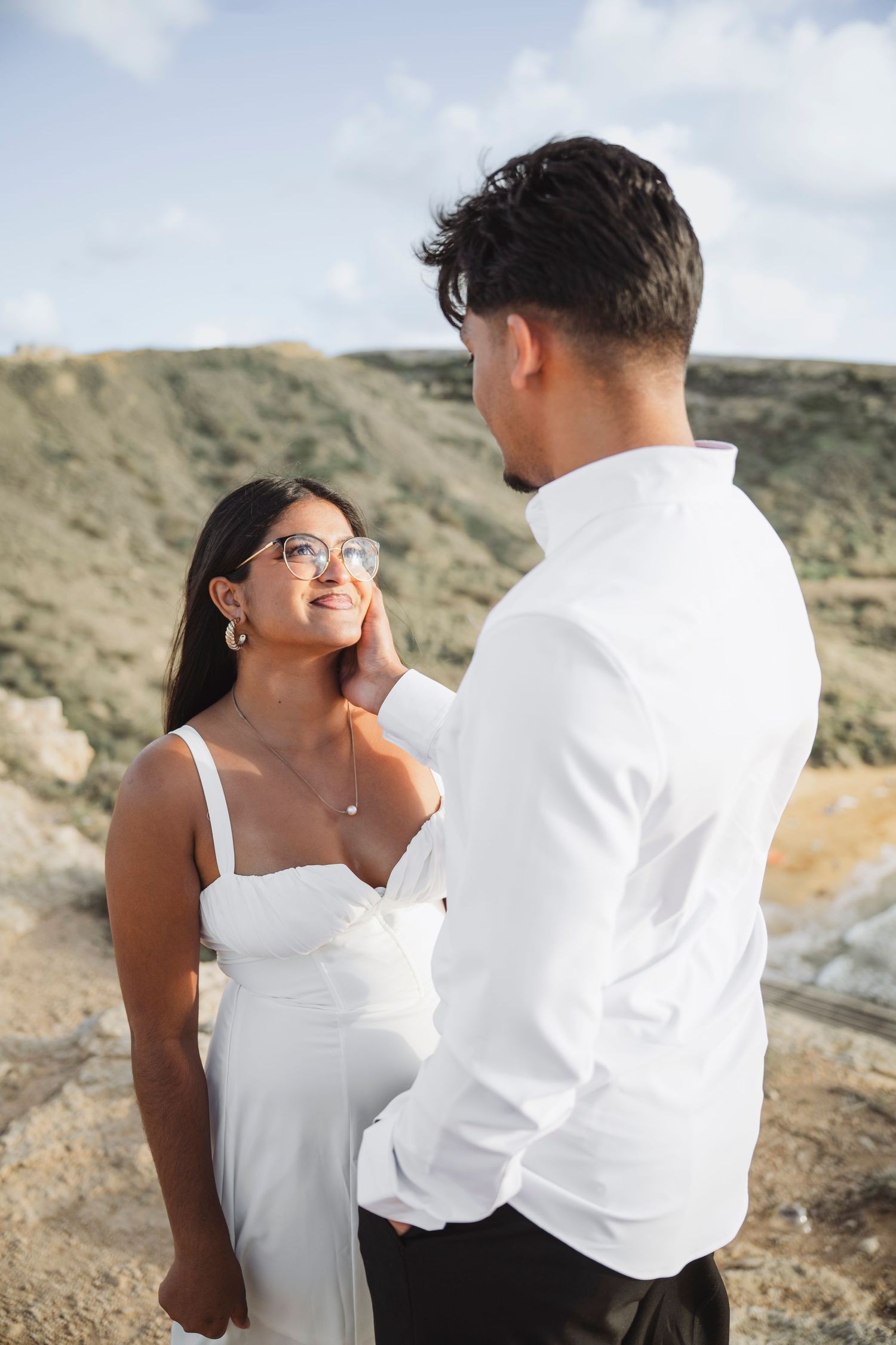 Romantic coastal photoshoot with couple in white outfits against rocky cliffside backdrop.