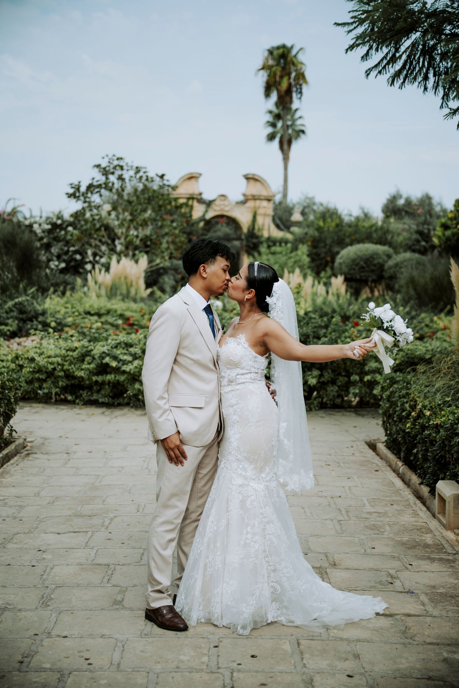Bride and groom share a kiss in a lush garden, bride in lace mermaid gown with veil, groom in beige suit.