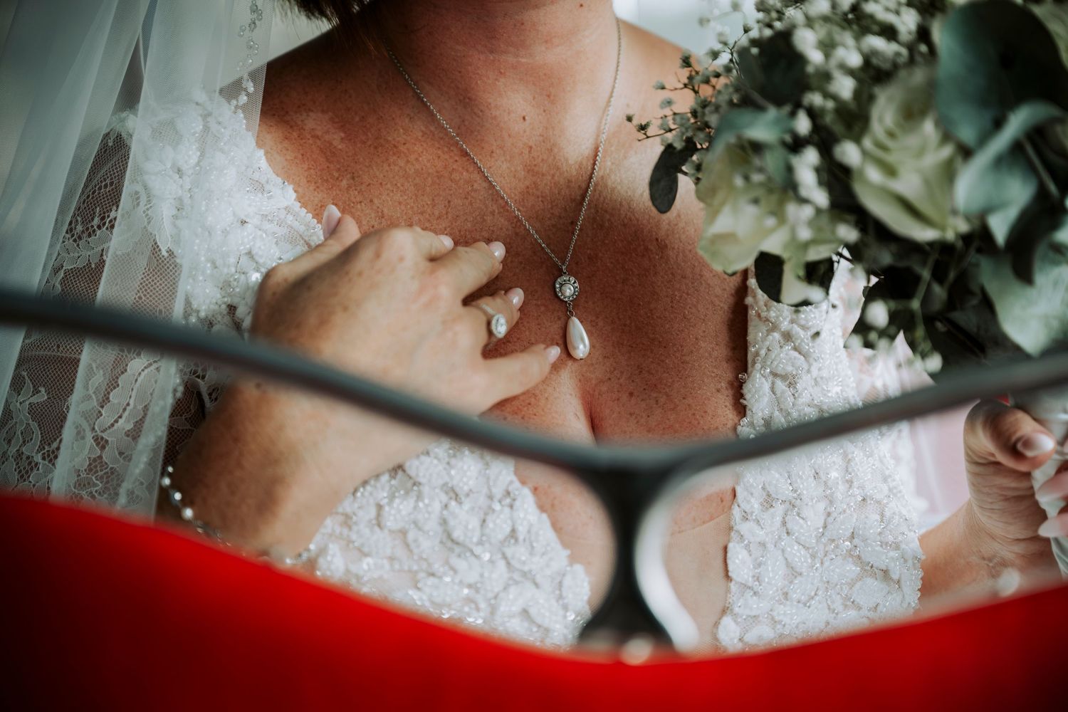 Close-up of a bride in a lace wedding dress wearing a teardrop necklace and ring, holding a bouquet of greenery.
