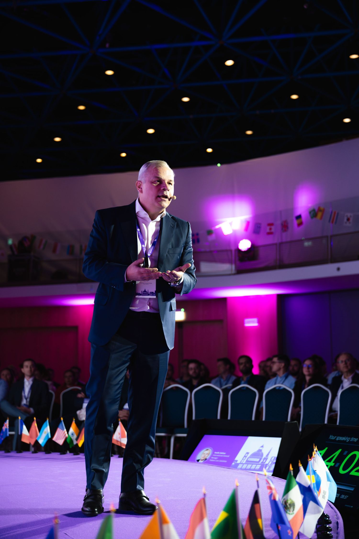 Professional speaker presenting on stage at corporate event with colorful lighting and national flags.