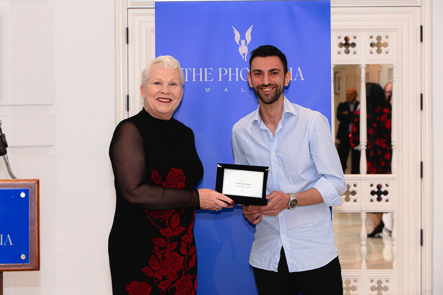 Award presentation ceremony against blue Phoenicia backdrop in elegant white interior setting.