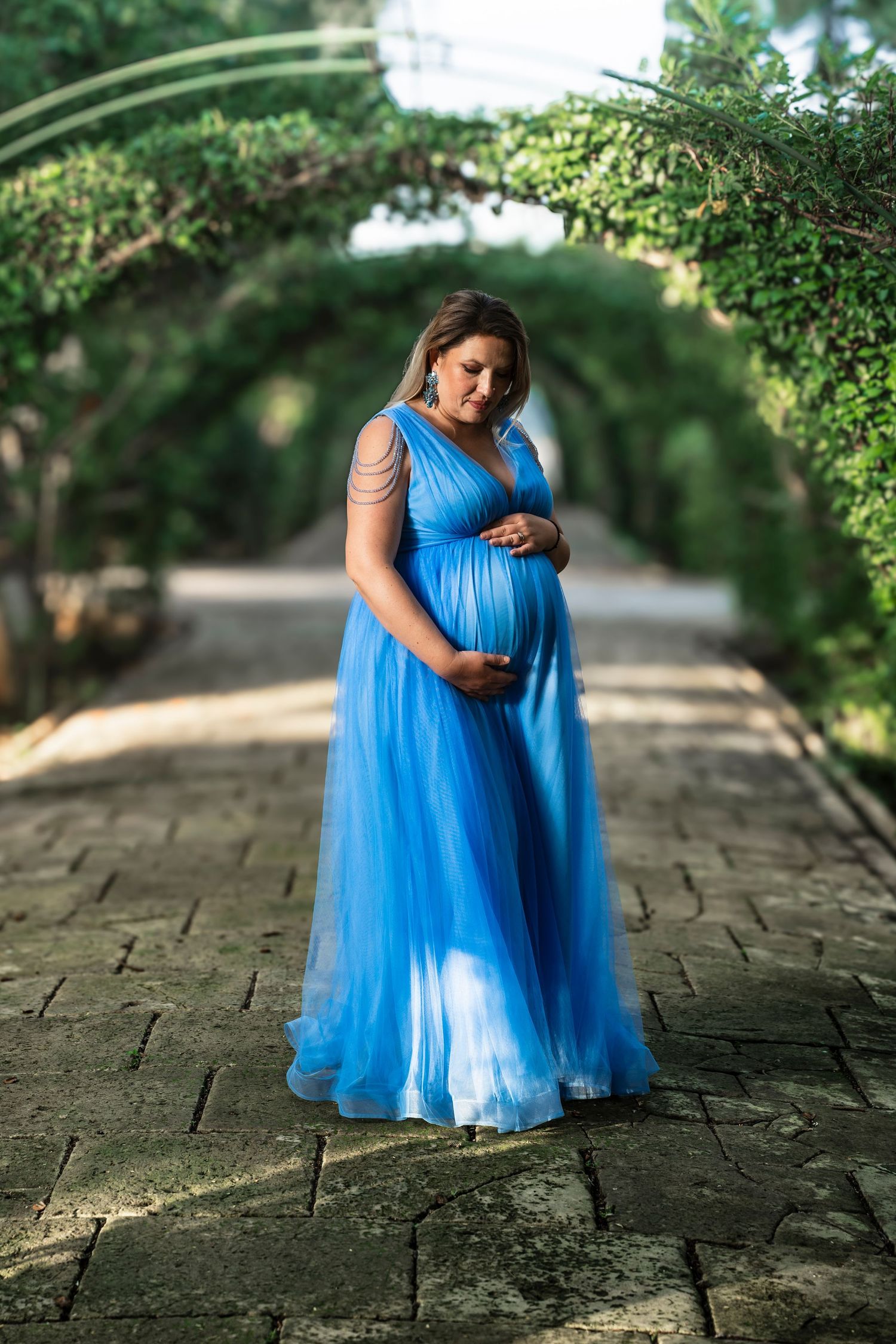 Pregnant woman in flowing blue tulle gown standing on stone path beneath lush green garden archway.