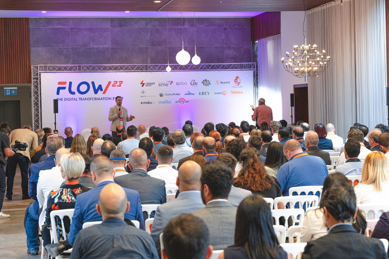 Large audience gathered in a modern conference room with purple lighting for the FLOW business event.