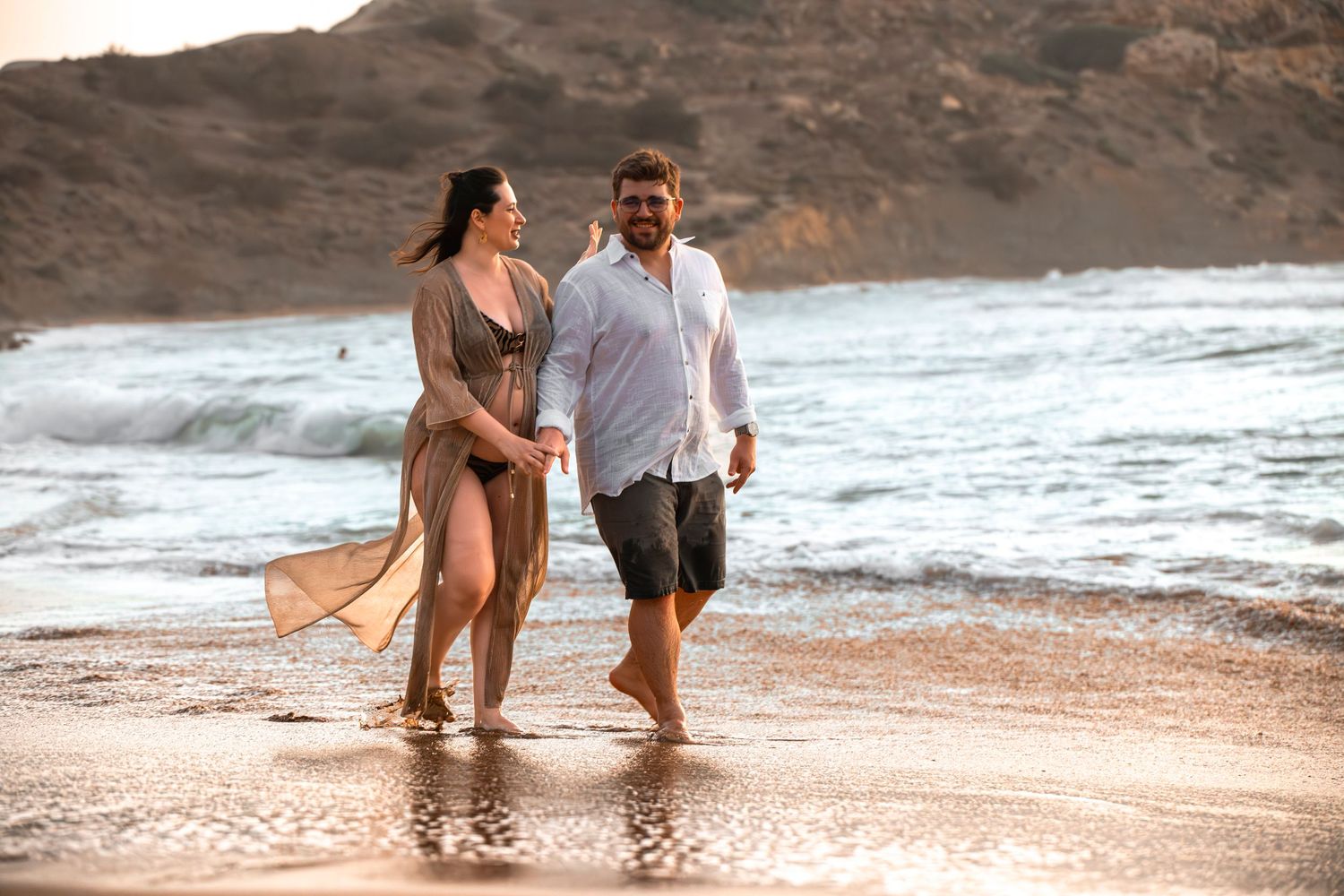 Couple walking hand in hand along a sandy beach at sunset, woman in bikini cover-up and man in white shirt.