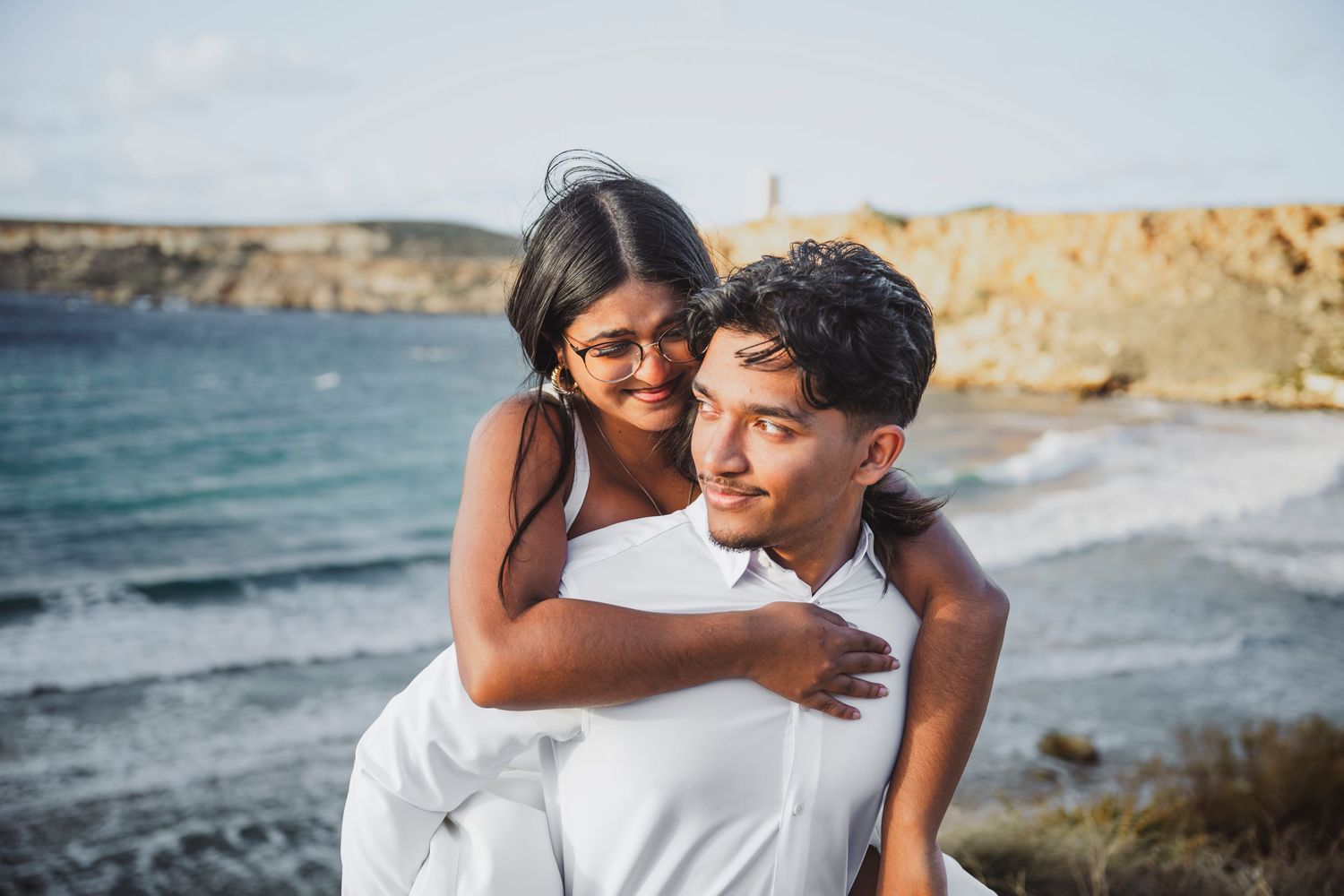 Couple in white clothing embracing on scenic beach with ocean views.