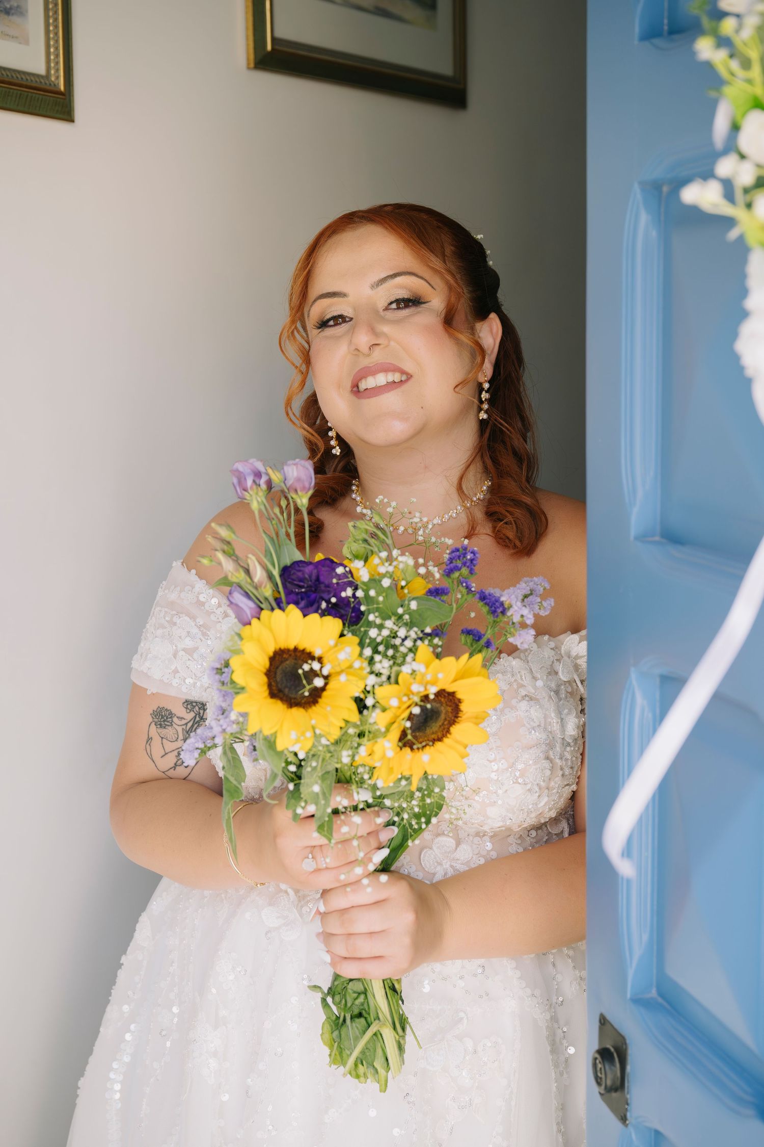 Smiling bride in white lace off-shoulder gown holding sunflower and purple bouquet beside a blue door.