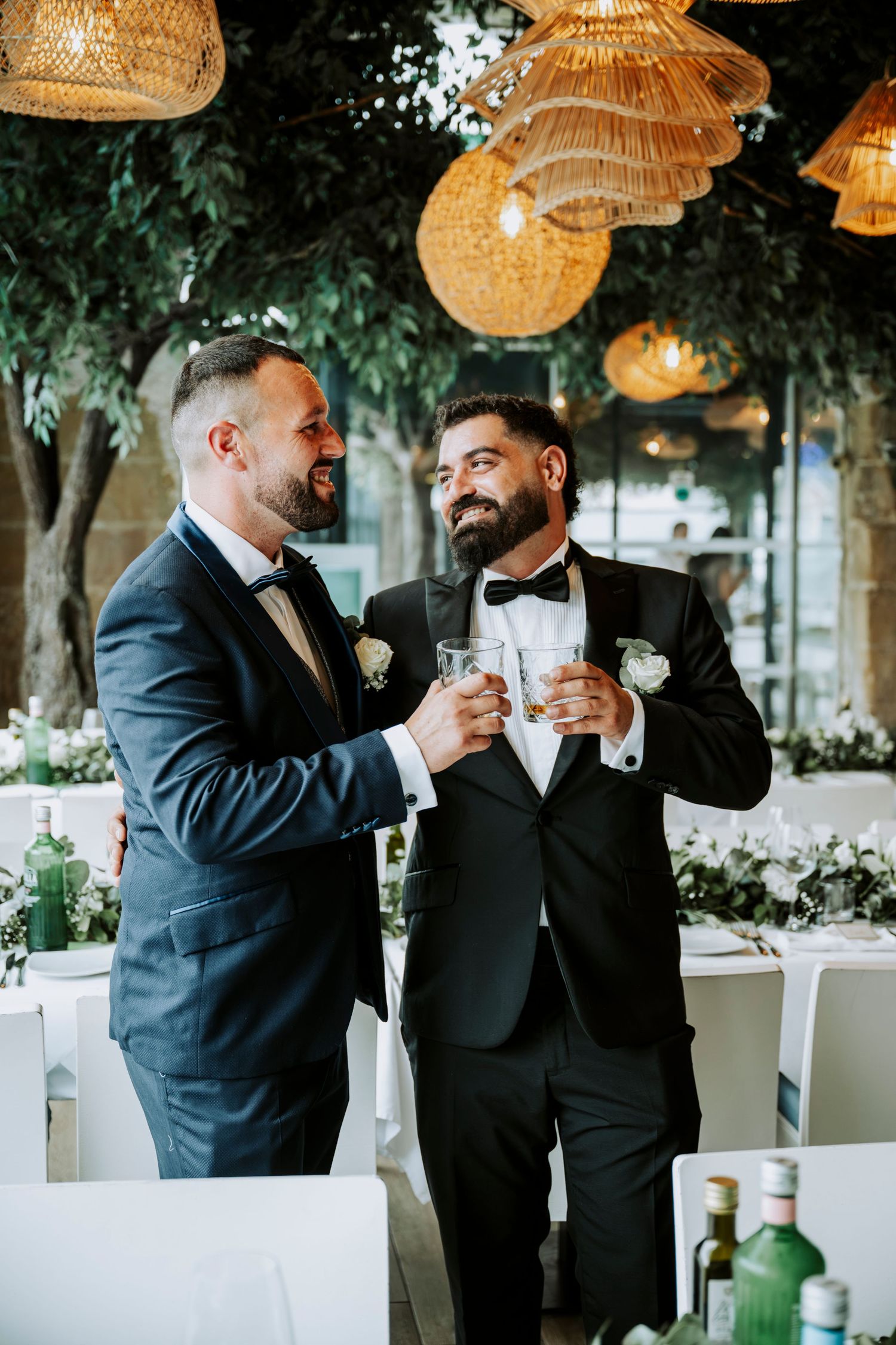 Two grooms in tuxedos toasting with drinks at their wedding reception under warm wicker pendant lights.