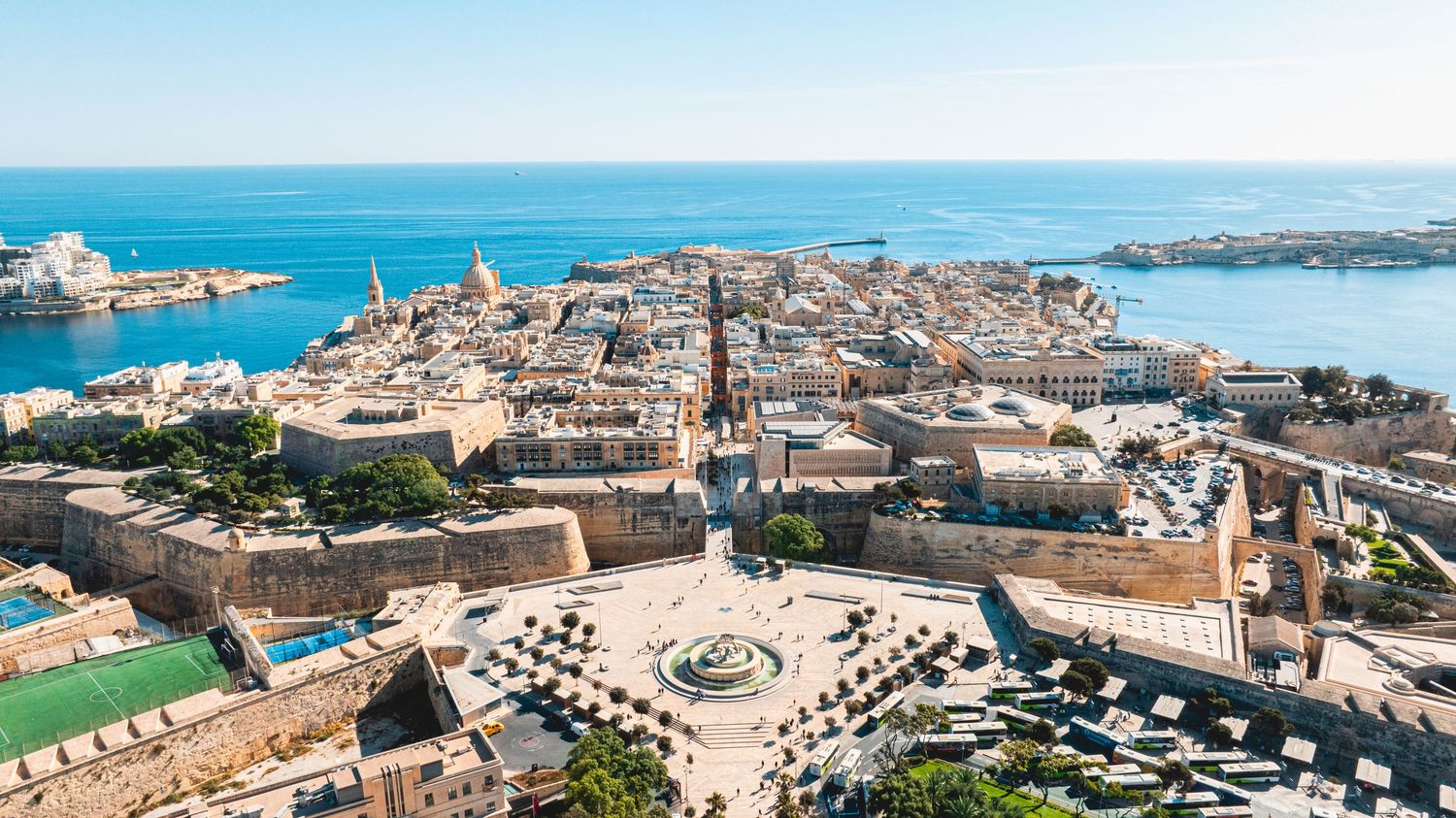 Sweeping aerial view of Malta's coastline showing urban development and historic architecture along blue waters.