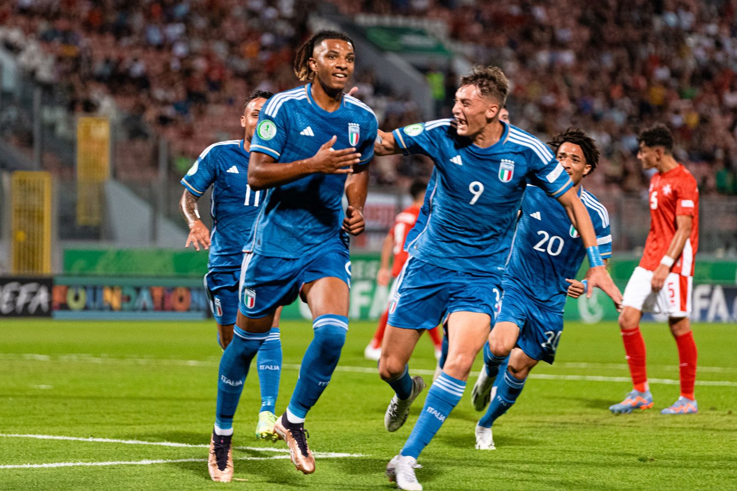 Soccer players in blue uniforms celebrate on field with stadium crowd in background.