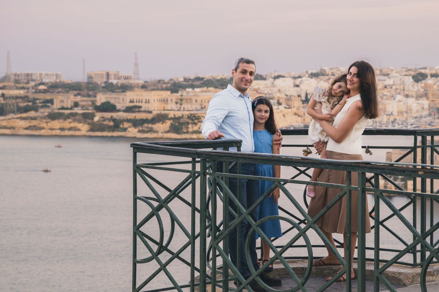 Family of four posing on a scenic balcony overlooking a Mediterranean coastal city at dusk.