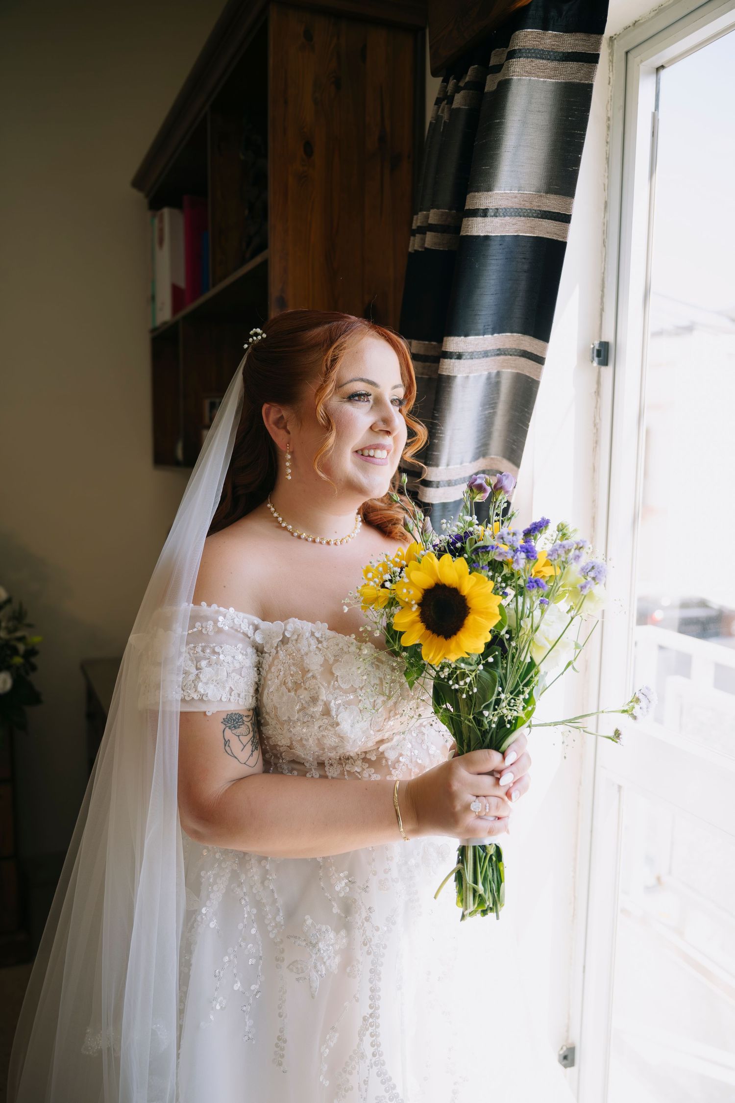 Smiling bride in off-shoulder lace gown holds sunflower bouquet by window, wearing pearl necklace and veil.