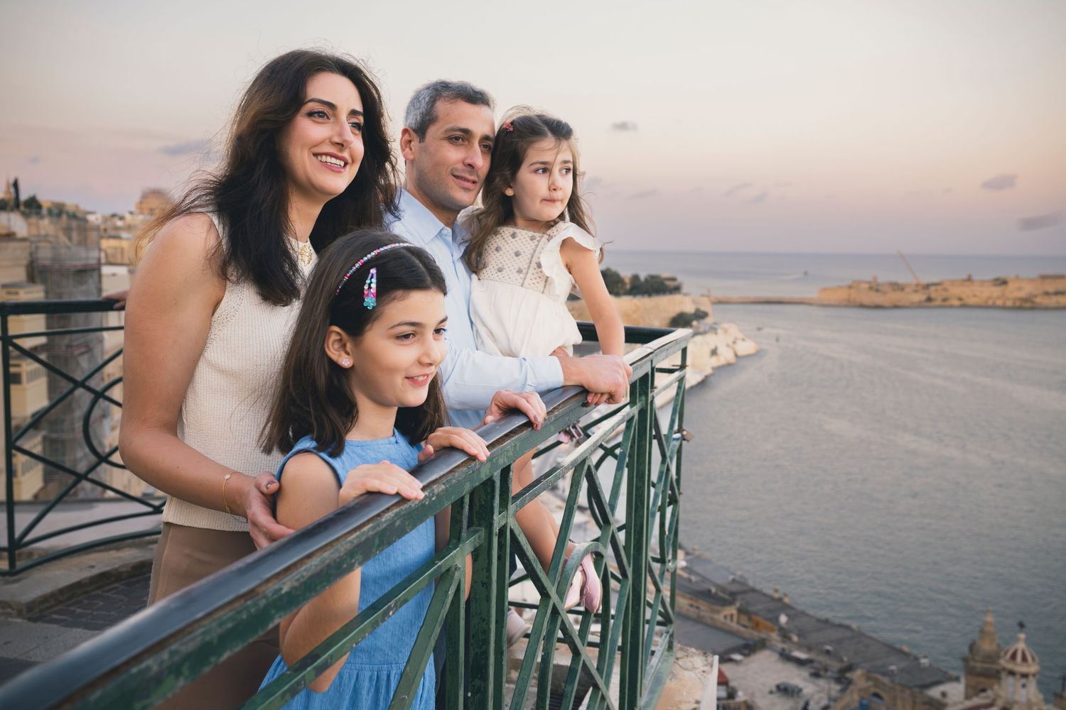 Family of four enjoying a scenic waterfront view from a balcony at sunset in Malta.