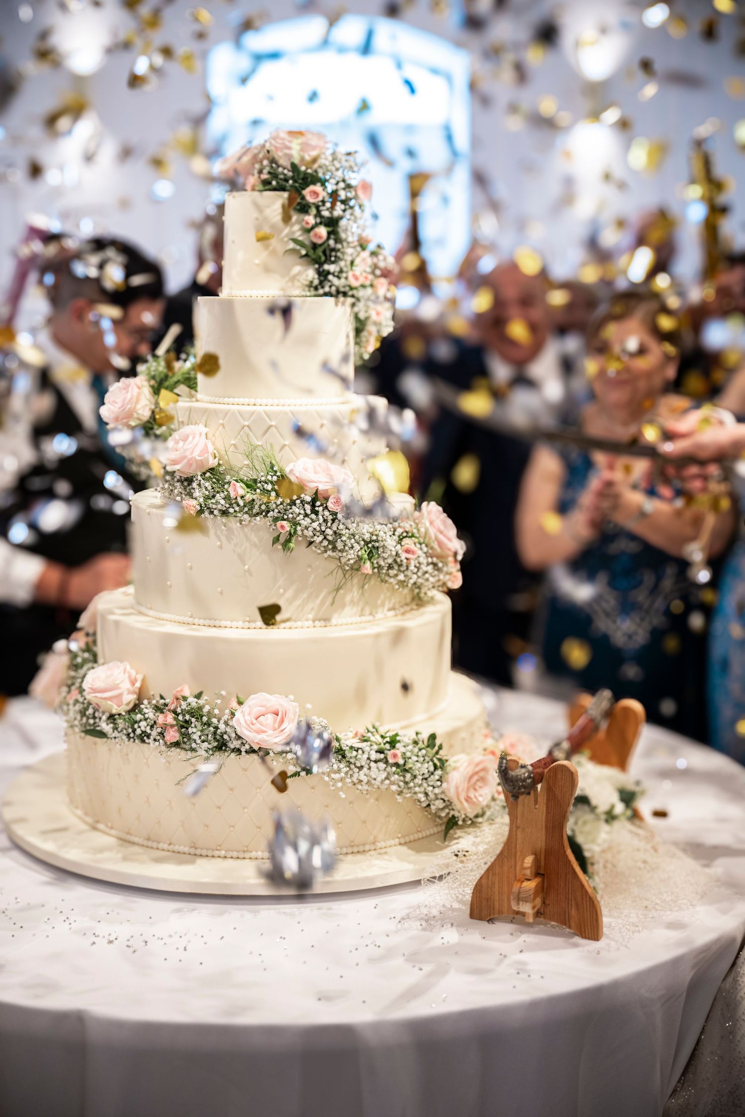 Elegant white tiered wedding cake decorated with delicate flowers and confetti falling in the background.