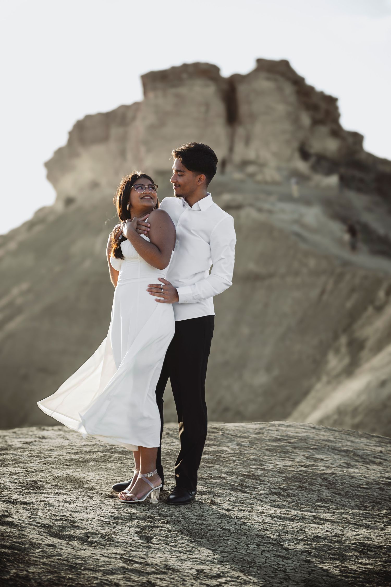 Romantic photoshoot on rocky cliff overlooking ocean with couple in matching white outfits.