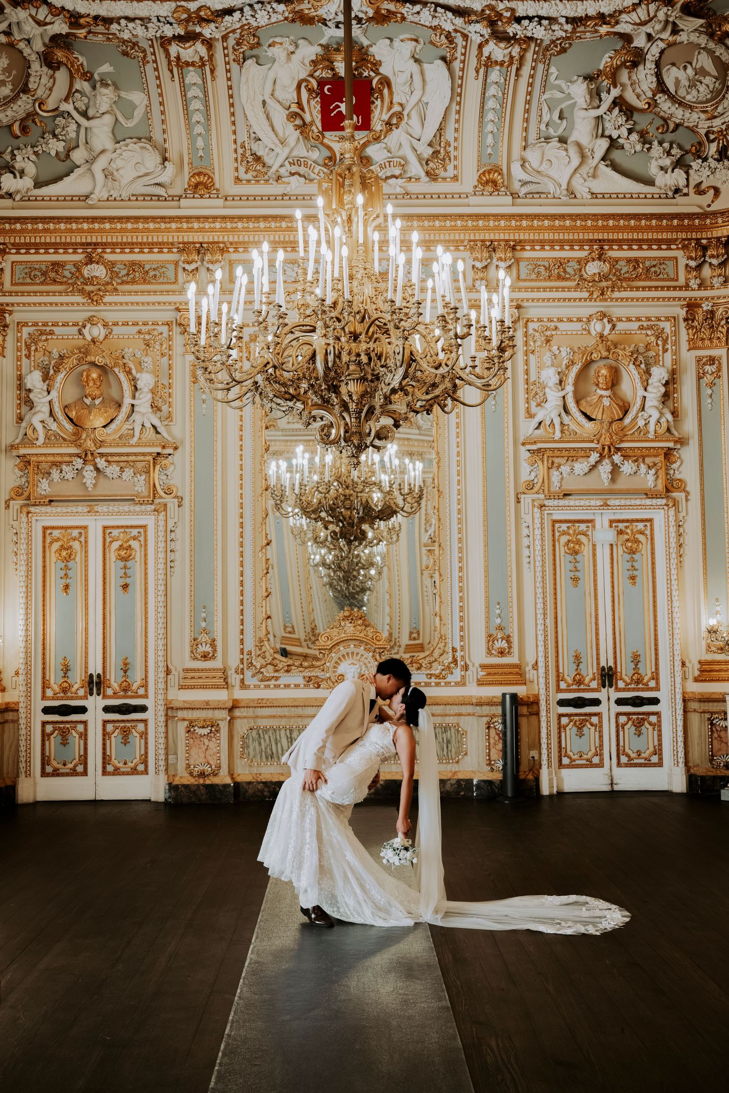 Bride and groom share a romantic dip kiss in an ornate golden ballroom with a grand chandelier.