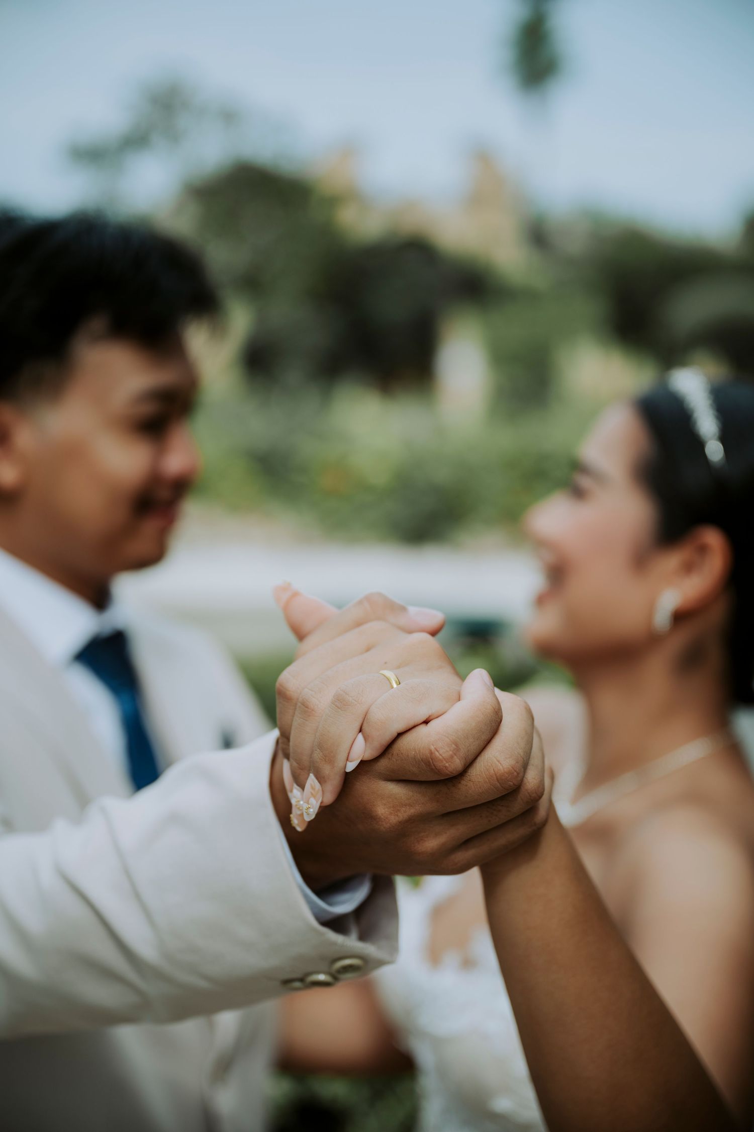 A bride and groom hold hands showing a wedding ring during their outdoor ceremony, smiling joyfully at each other.