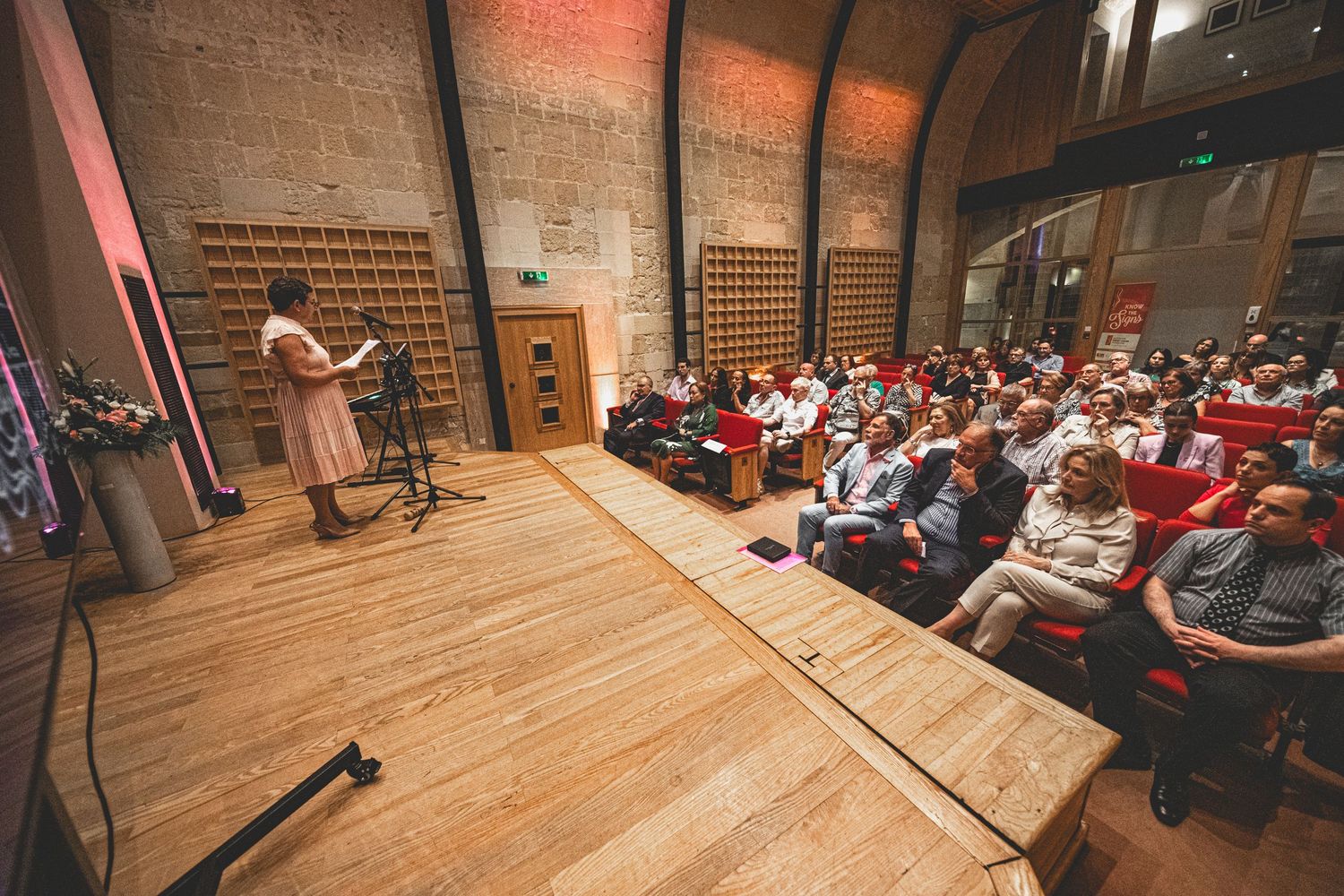Speaker presenting to a full audience in an auditorium with wooden stage and brick walls.