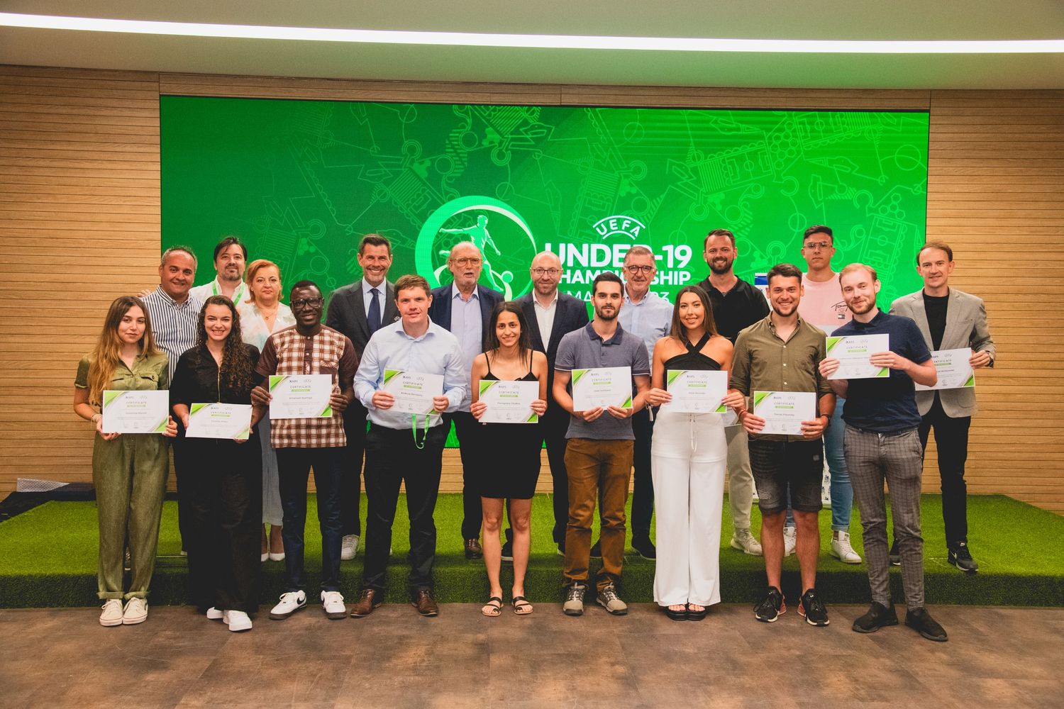 Group of award recipients standing in line holding certificates against a green branded backdrop.