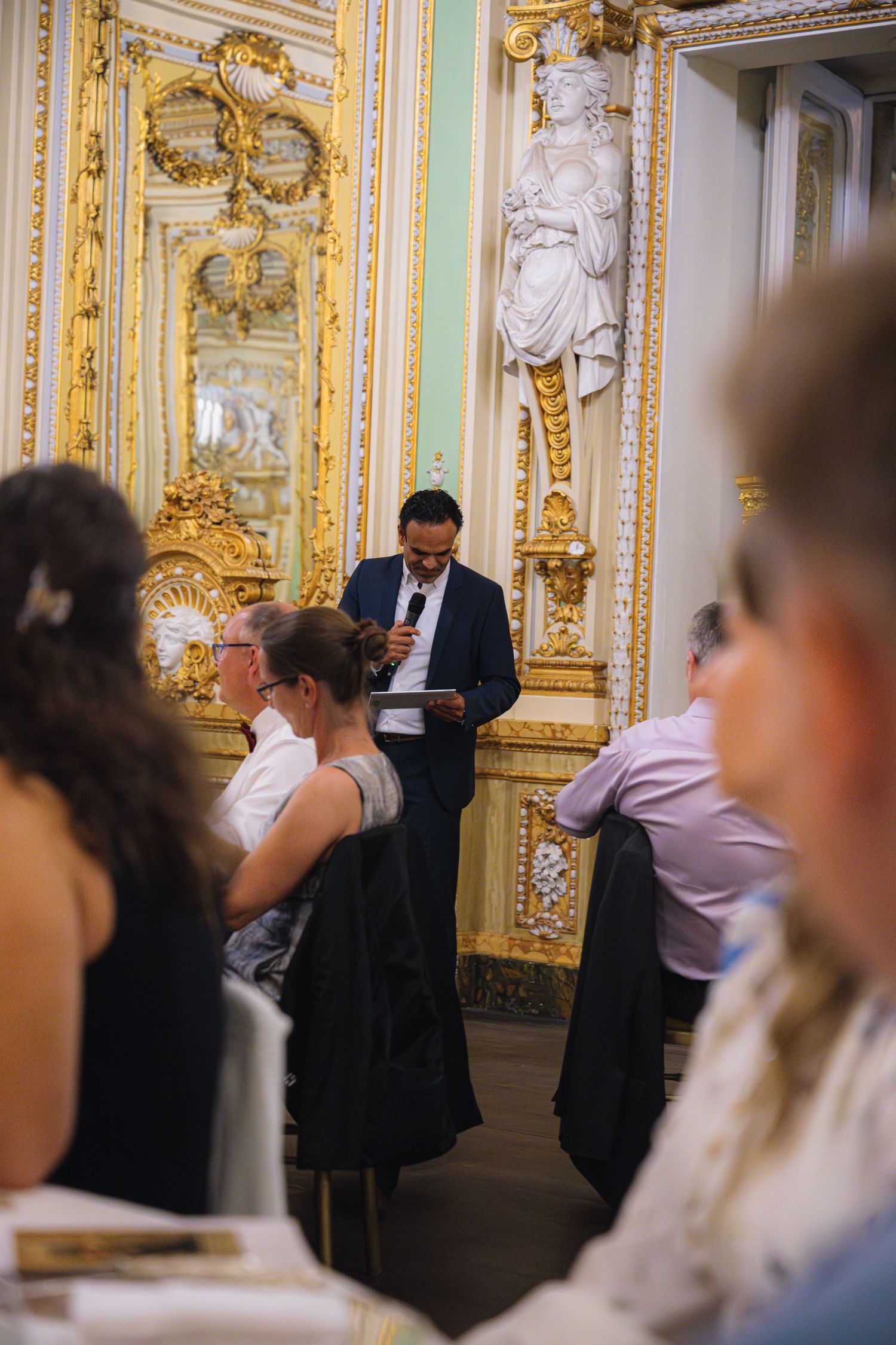 Formal dinner gathering in an ornate baroque-style room with gold mirrors and decorative moldings.