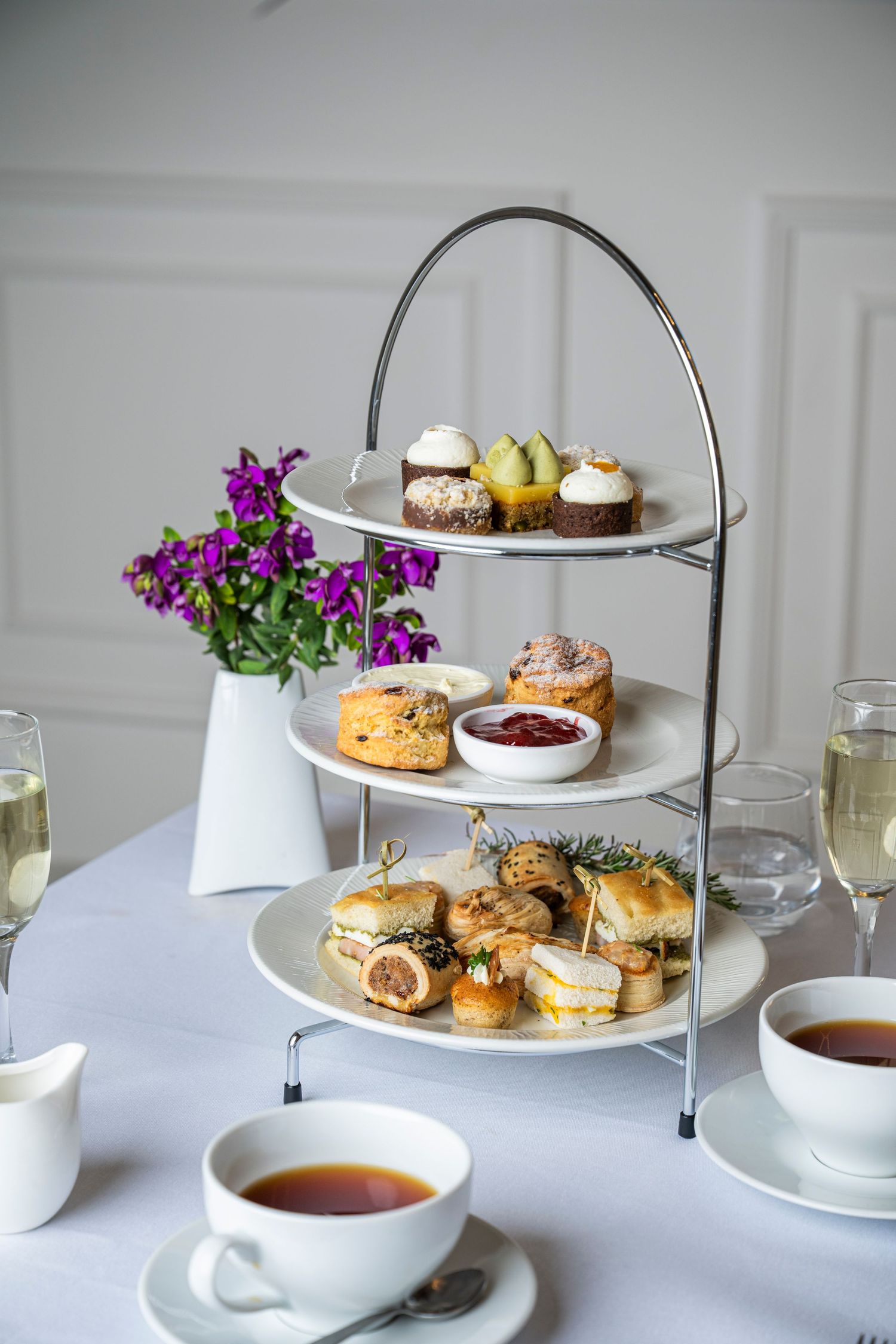 Elegant three-tier afternoon tea stand with sandwiches, scones, and pastries beside tea cups and champagne glasses.