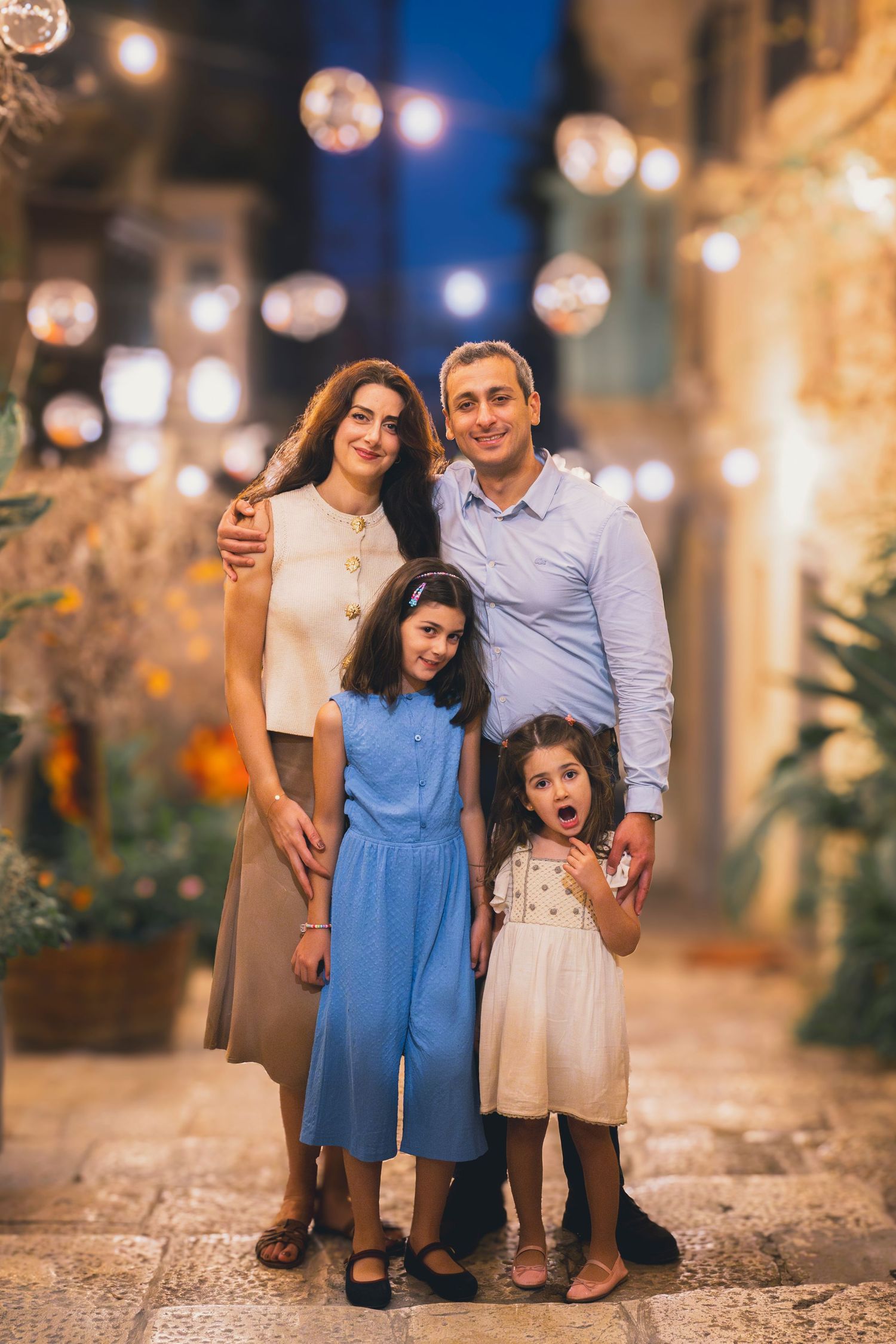 Family of four posing together on a charming cobblestone street at night with glowing string lights.
