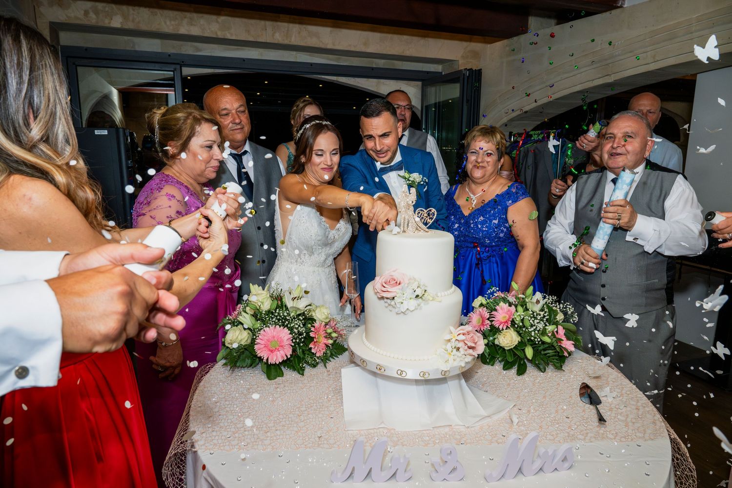 Wedding guests celebrate and react with joy as the newlyweds cut into their elegant white wedding cake.