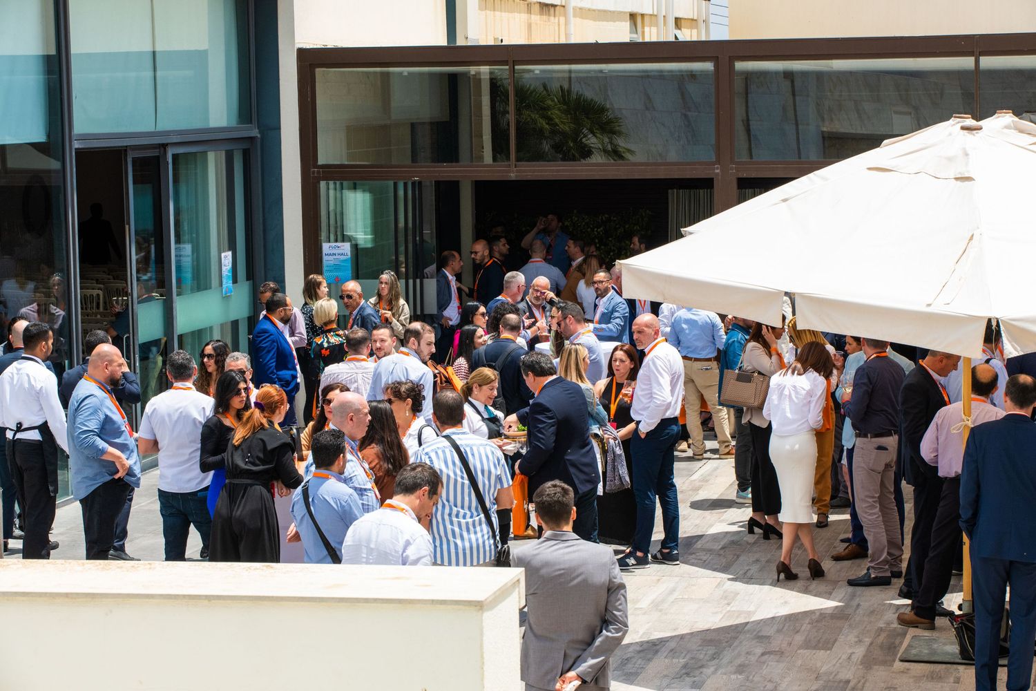 Large group of business people mingling during an outdoor networking event under white umbrellas.