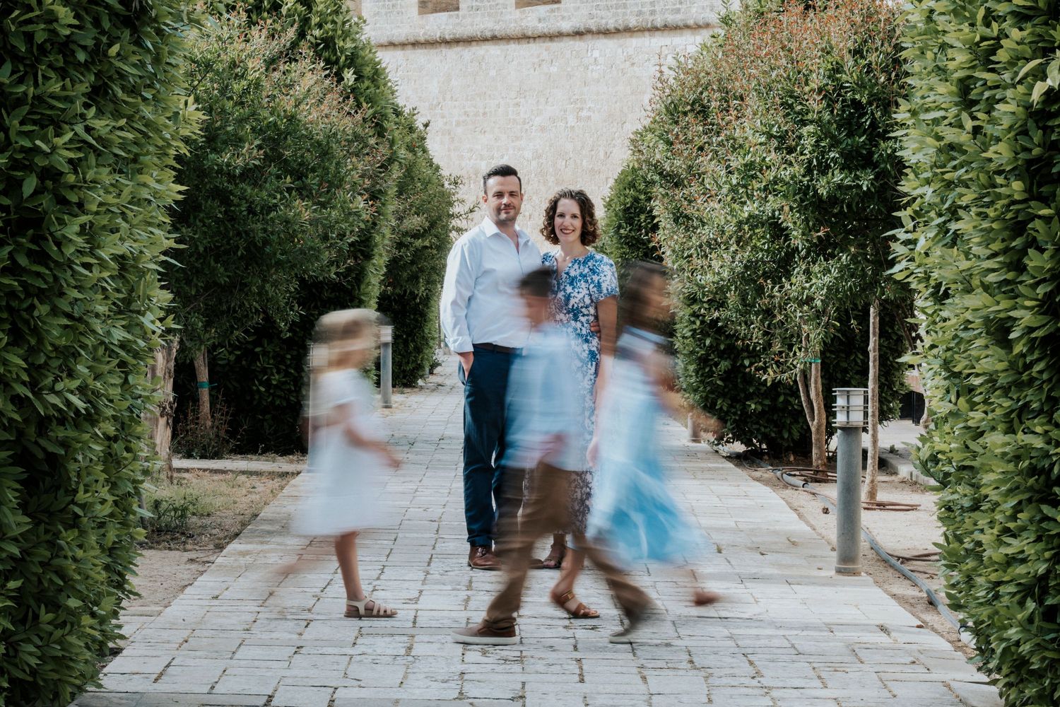 Family portrait of parents standing still while children run around them in a garden pathway lined with tall hedges.