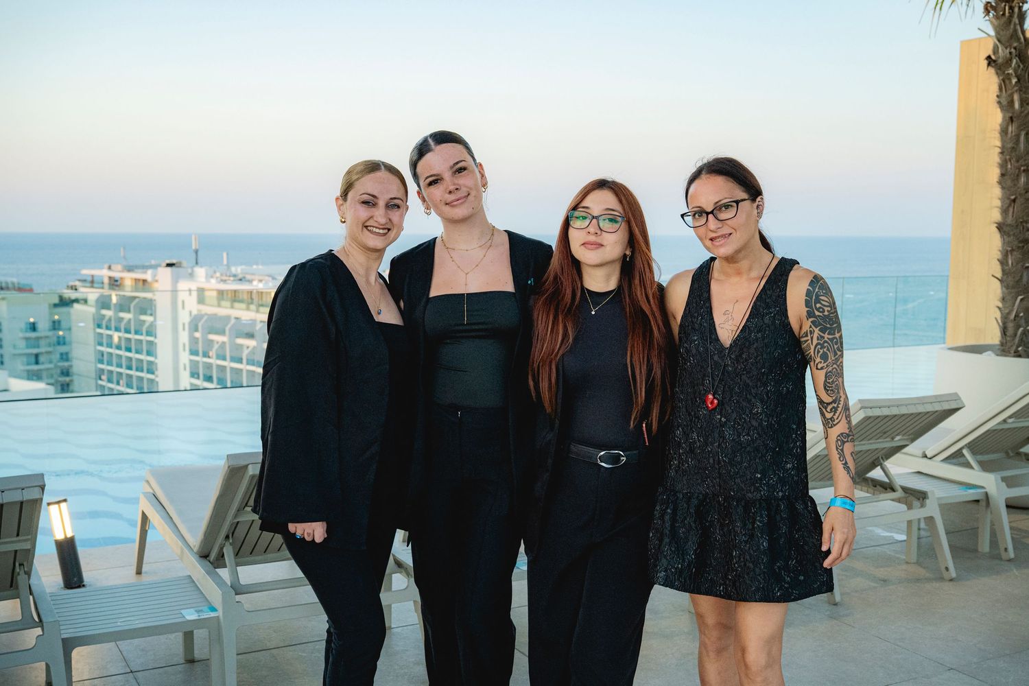 Four women in black outfits pose together on a rooftop terrace with ocean and city views in the background.