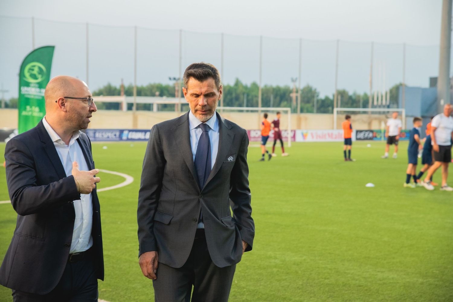 Two men in business attire having a discussion on a sports field during daytime.