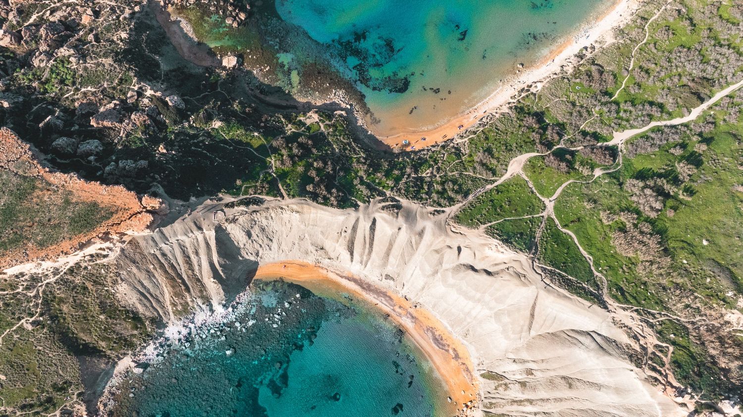 Sweeping coastal landscape with sandy coves, rocky cliffs and emerald waters viewed from above.