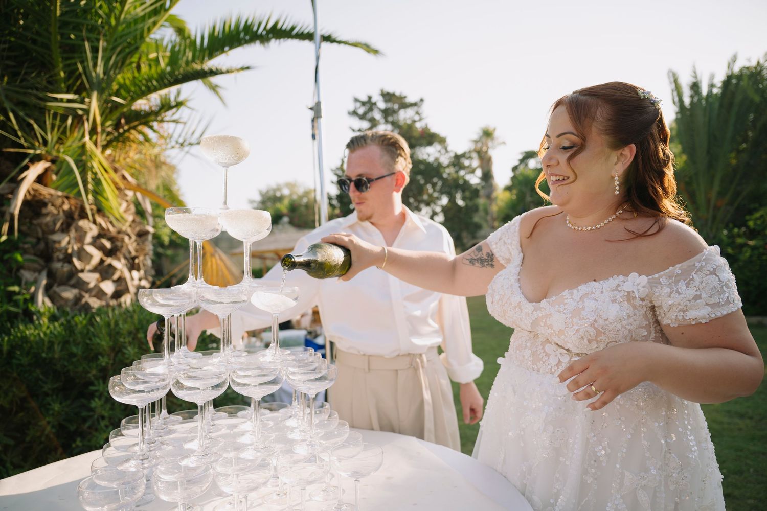 Bride and groom pouring champagne tower at outdoor wedding celebration surrounded by lush greenery.