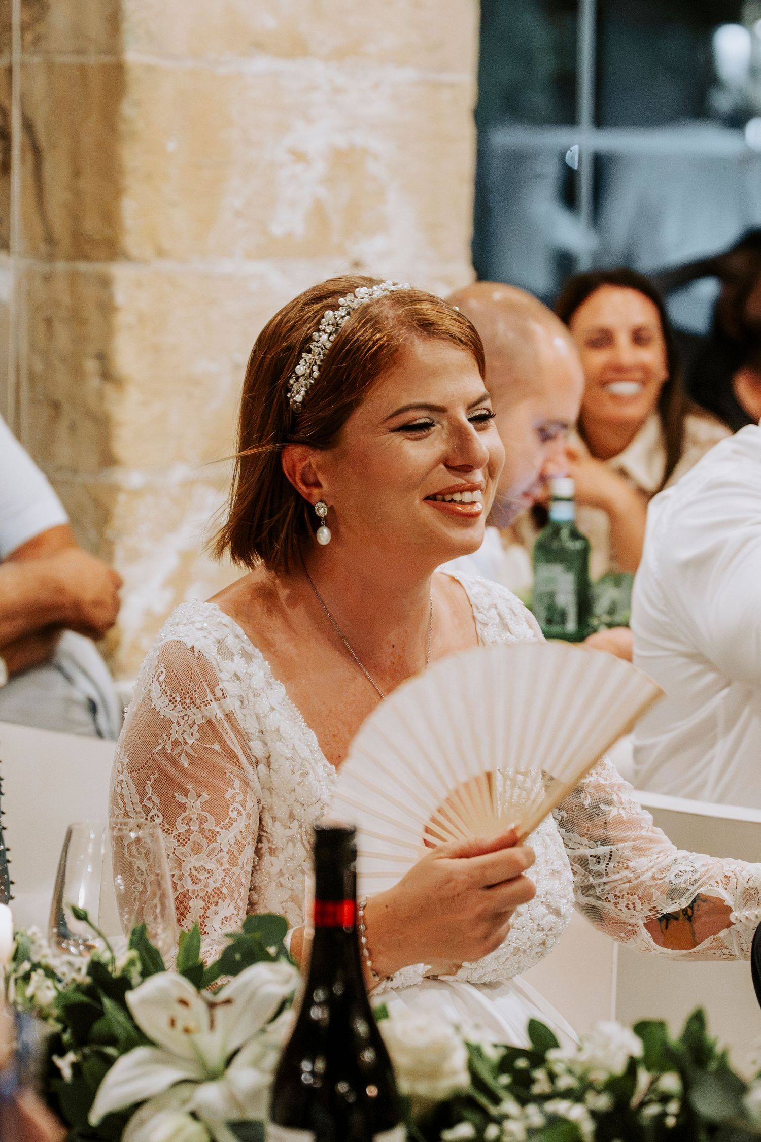 Smiling bride in lace dress holding a white hand fan at wedding reception with floral arrangements.