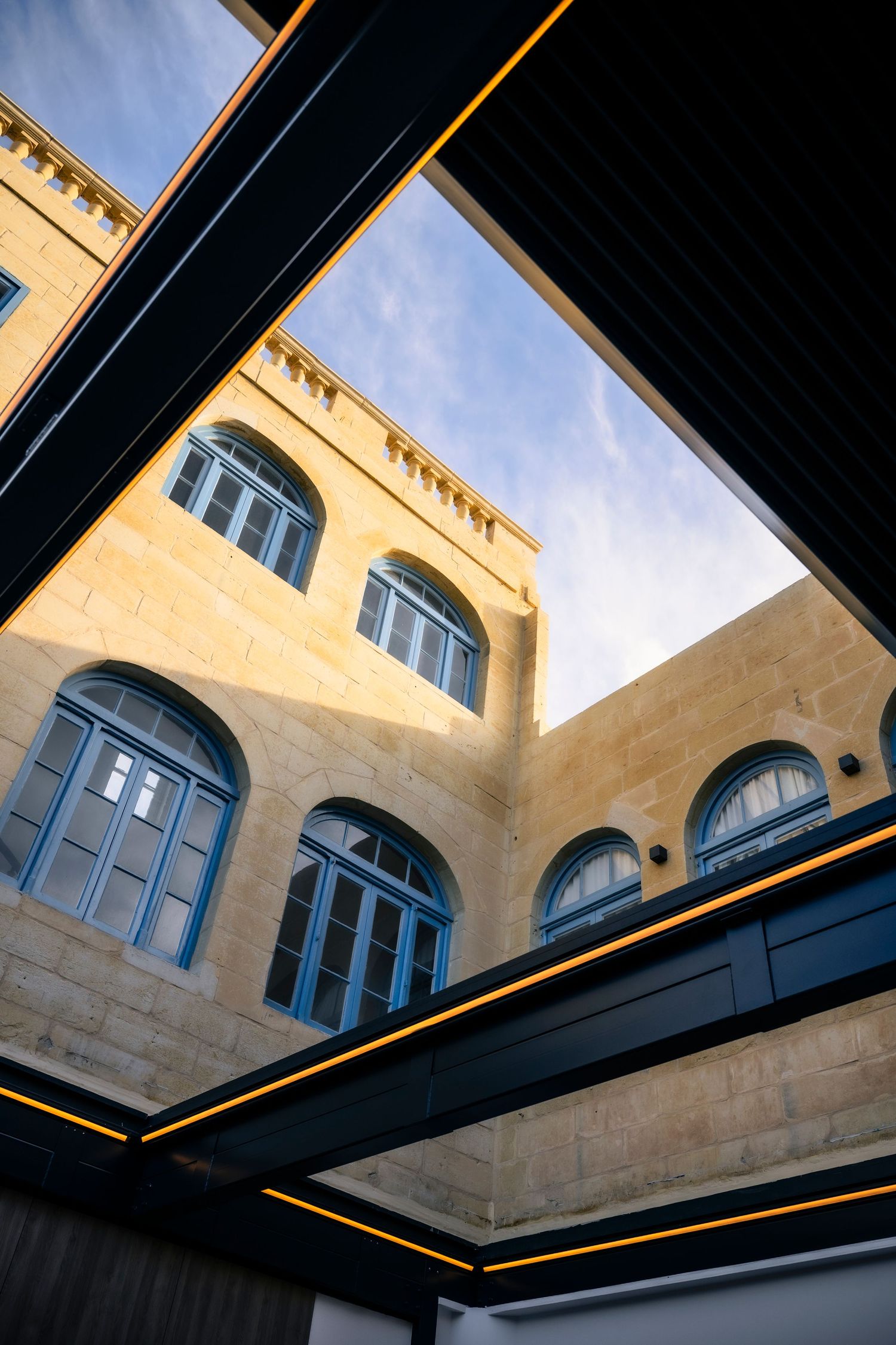 Looking up at sandstone building architecture with black beams and arched windows against blue sky.