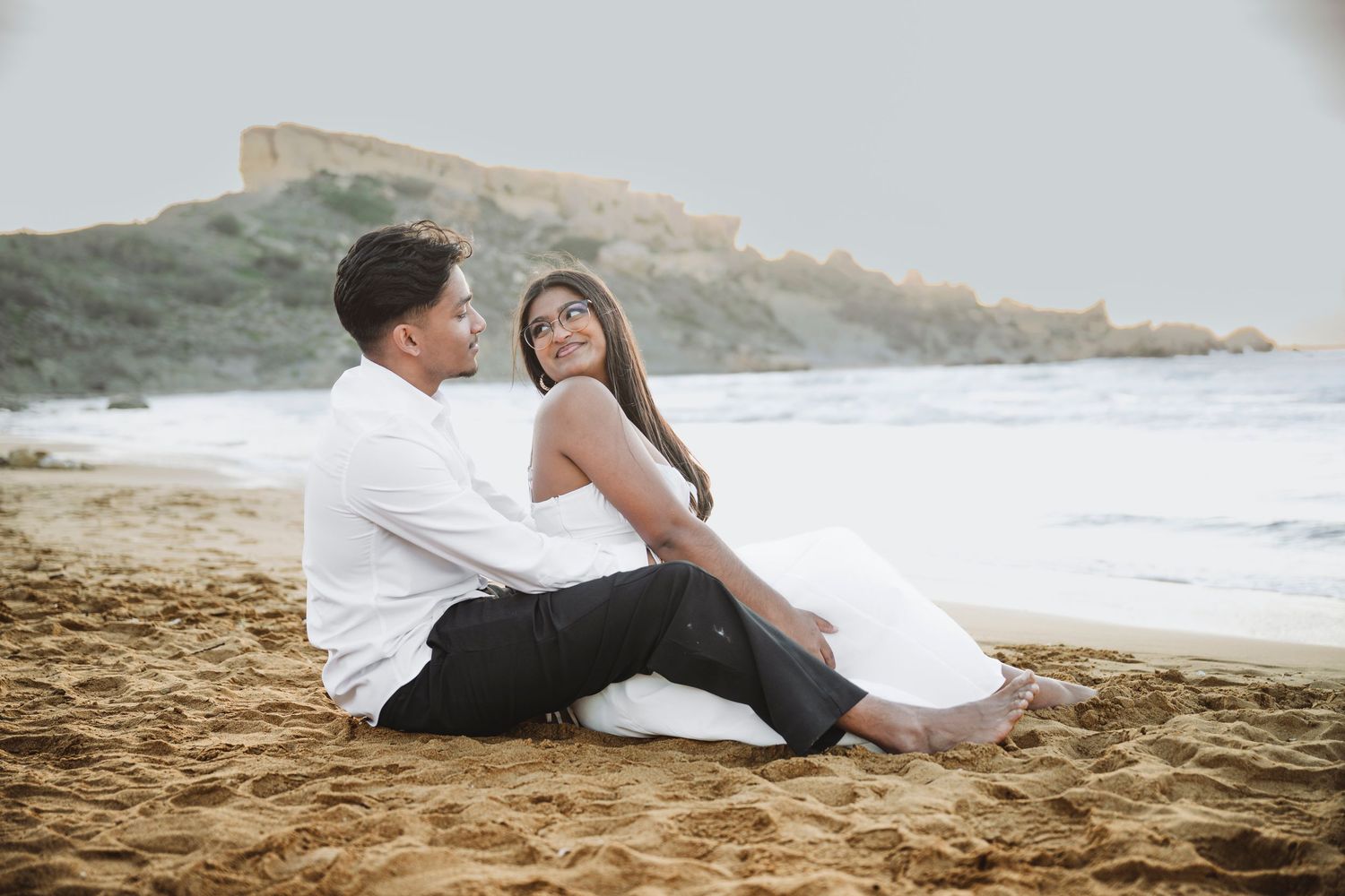 Couple relaxing on sandy beach with dramatic coastal cliffs in background.
