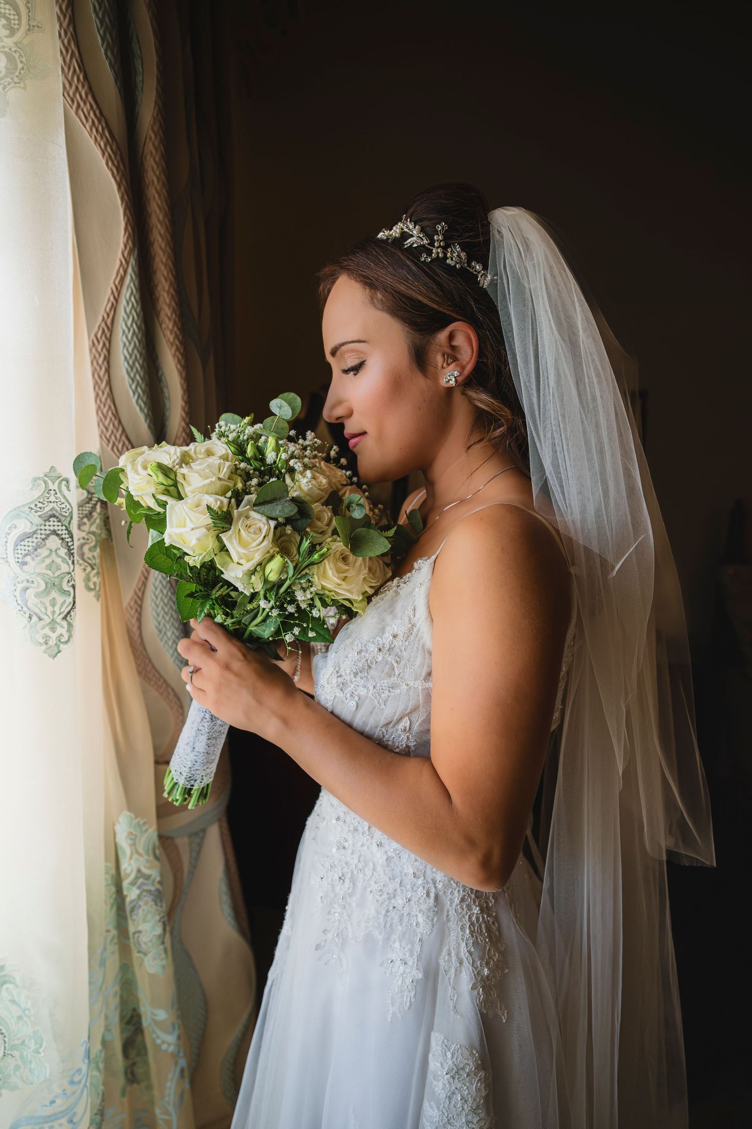 Bride in white lace dress holding and smelling a bouquet of white flowers near window.