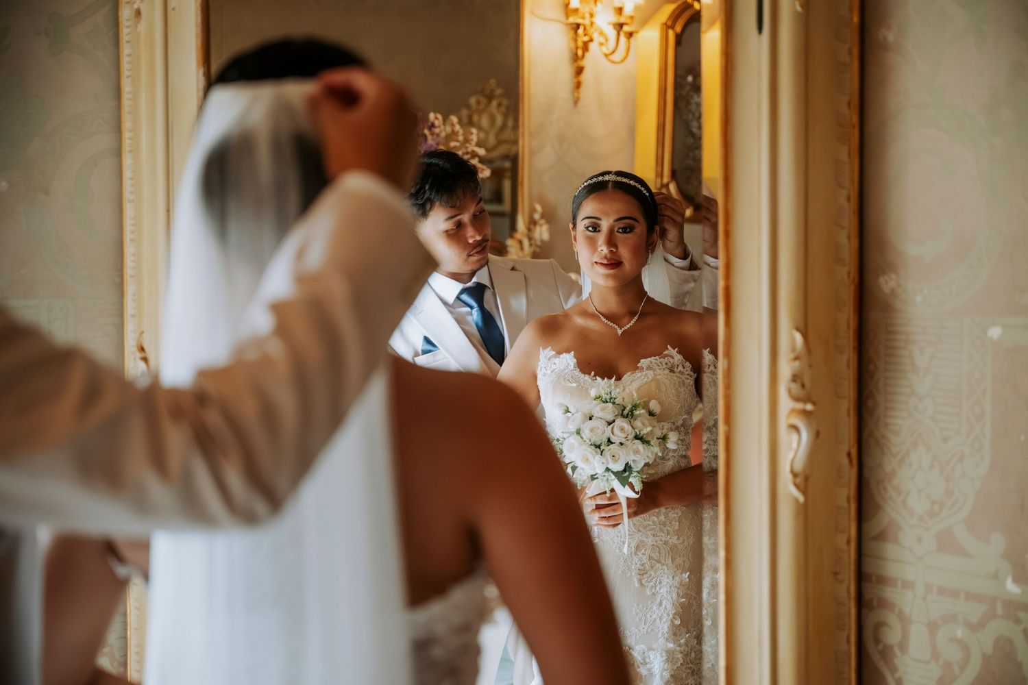 Bride in white gown holds bouquet while groom adjusts her veil, reflected in ornate gold mirror.
