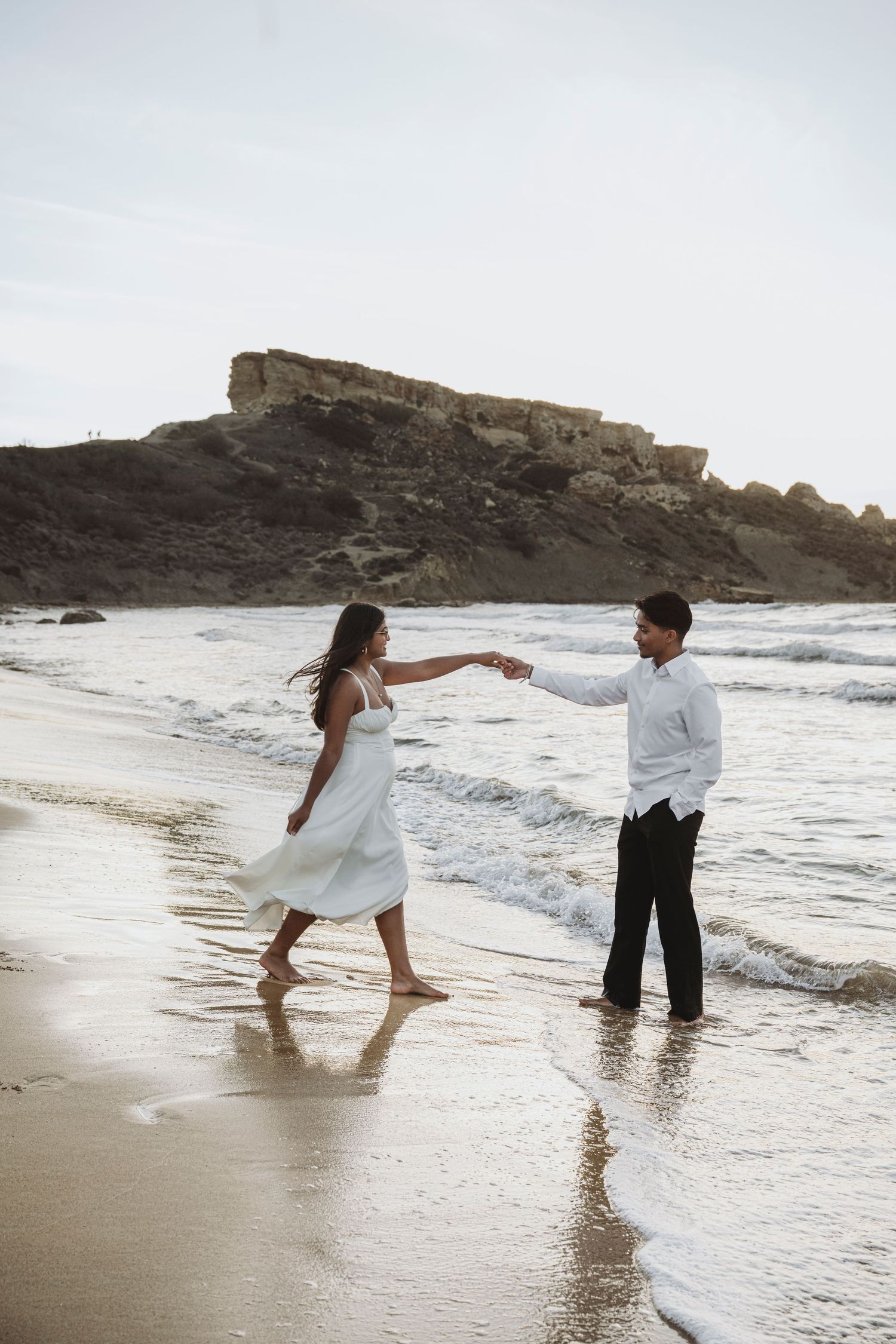 Playful couple splashing in ocean waves during beach photoshoot at sunset.