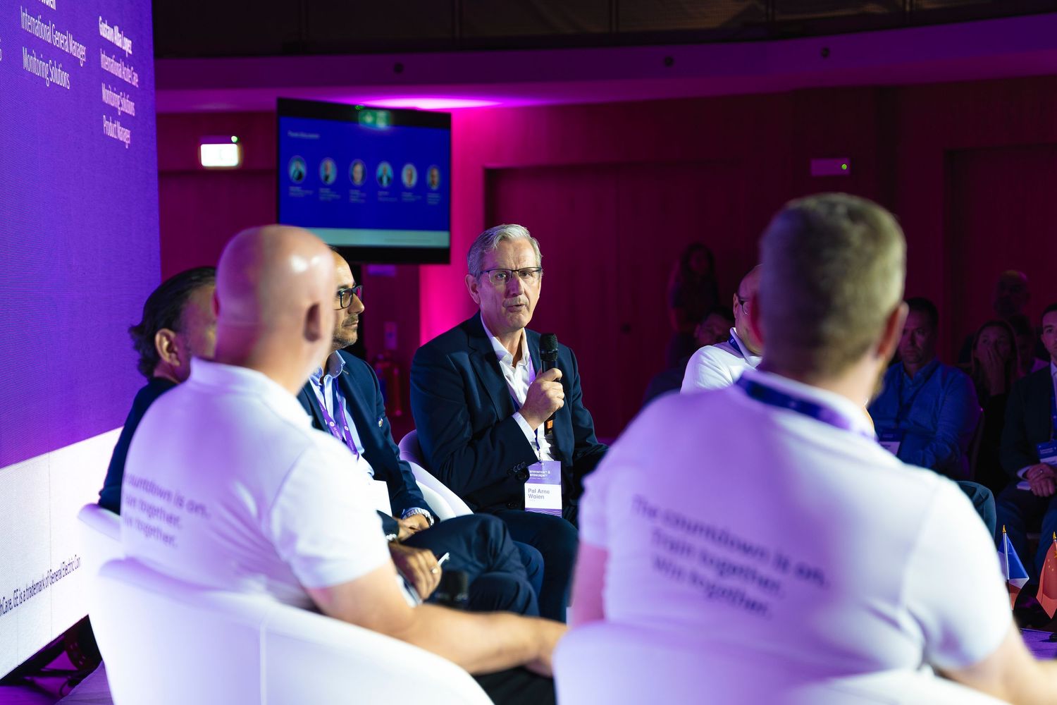 Conference attendees seated in rows with purple ambient lighting during presentation.