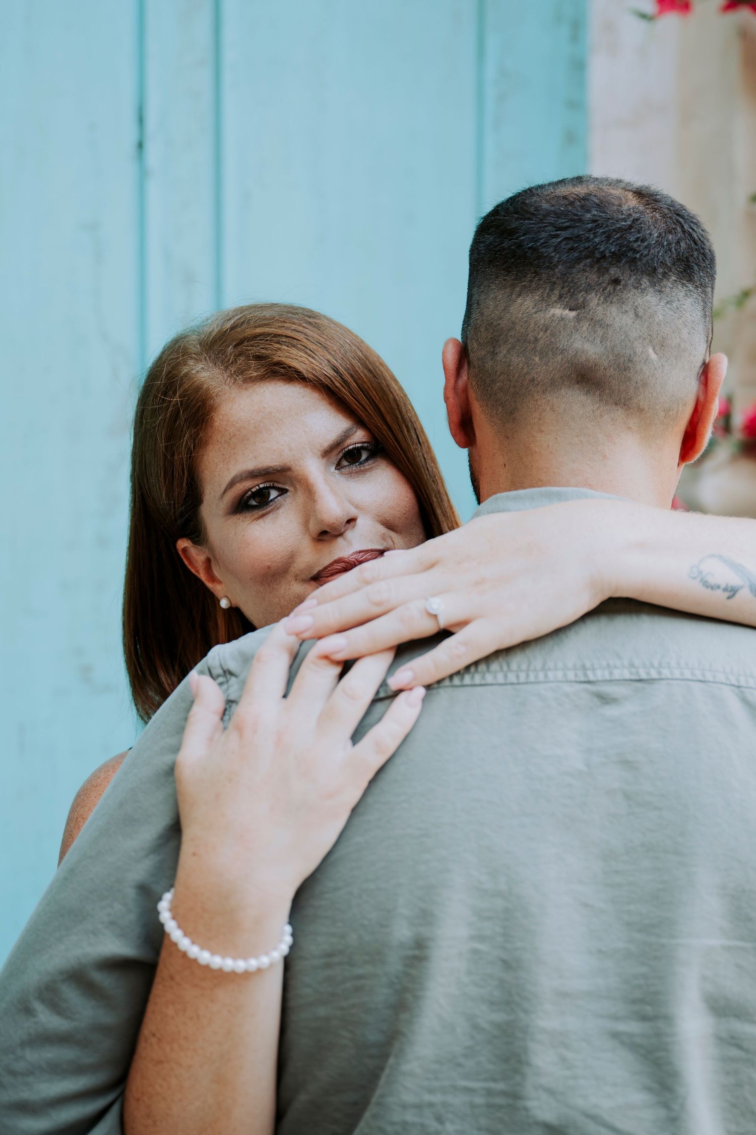 Woman with red hair embraces man from behind, showing off engagement ring, pearl bracelet, and tattoos against blue wall.