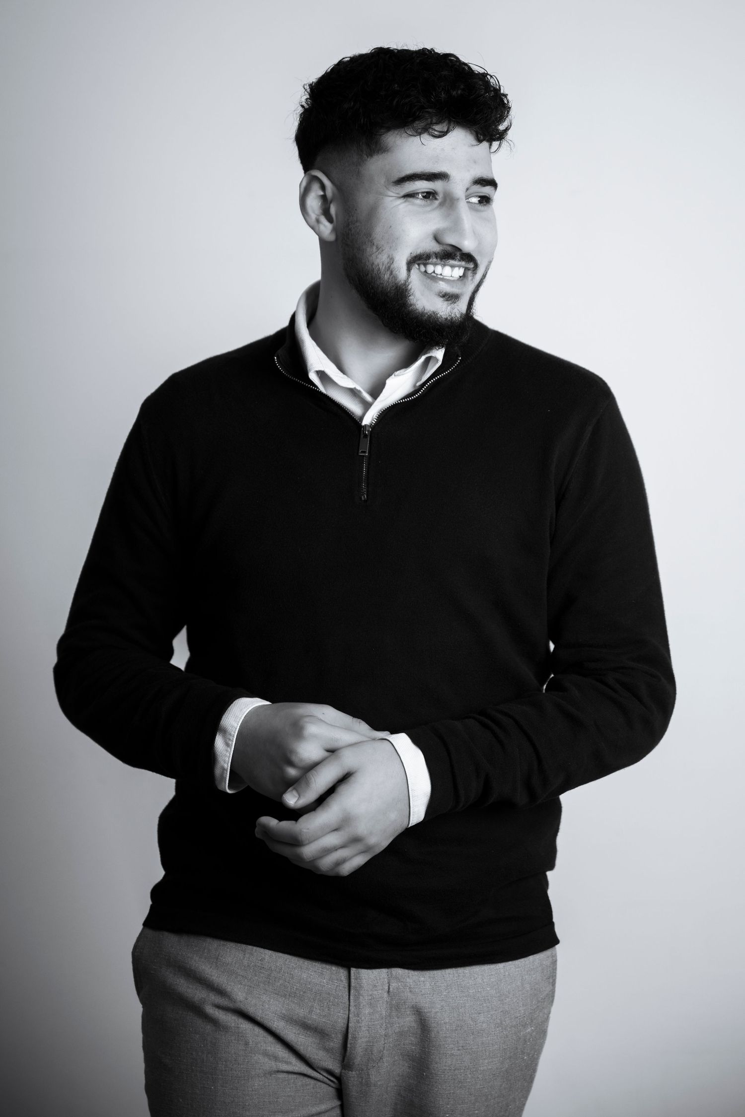 A person wearing a black sweater and gray pants poses against a white background in a casual portrait series.