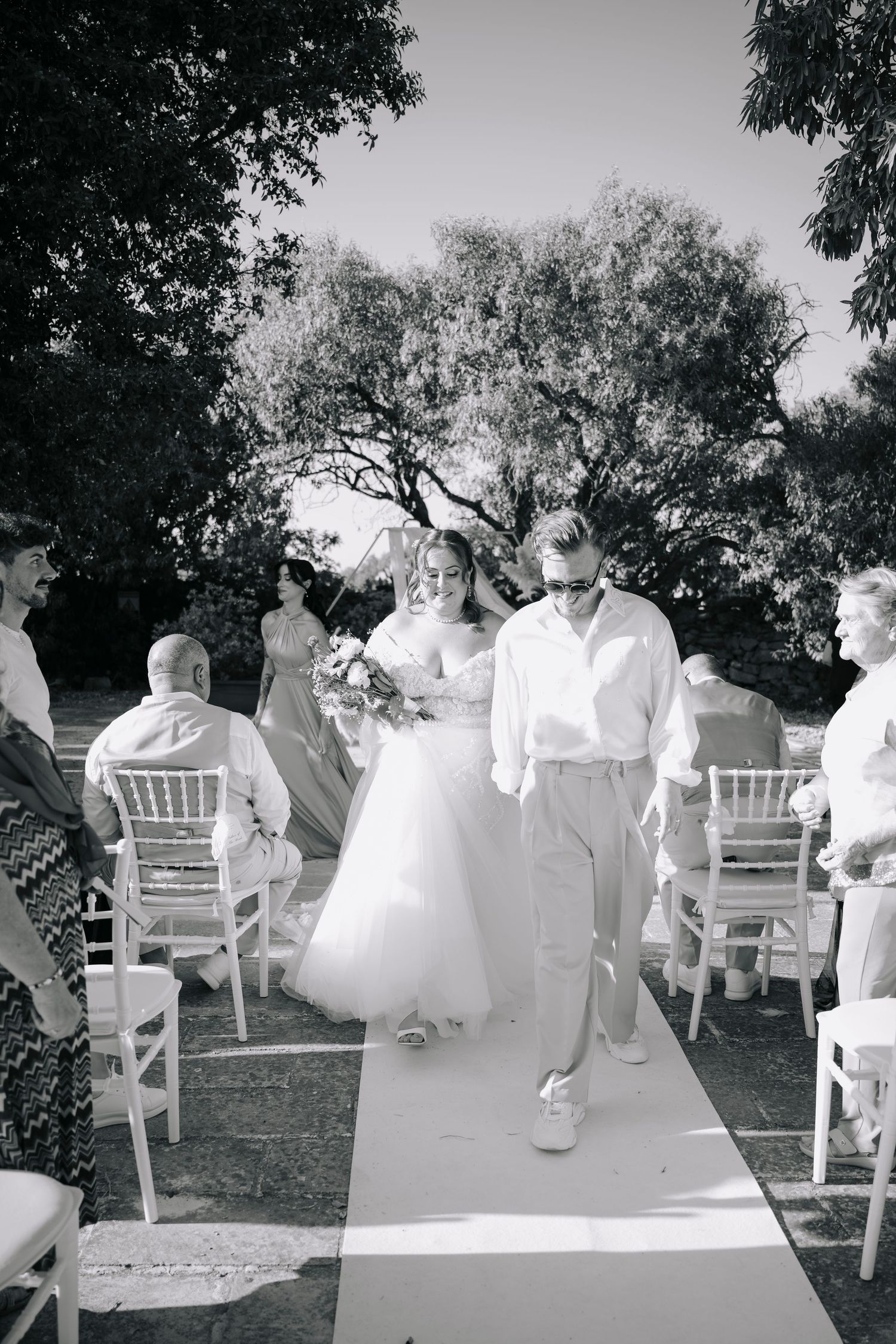 Black and white photo of a bride and groom walking down the aisle at an outdoor wedding ceremony surrounded by guests.