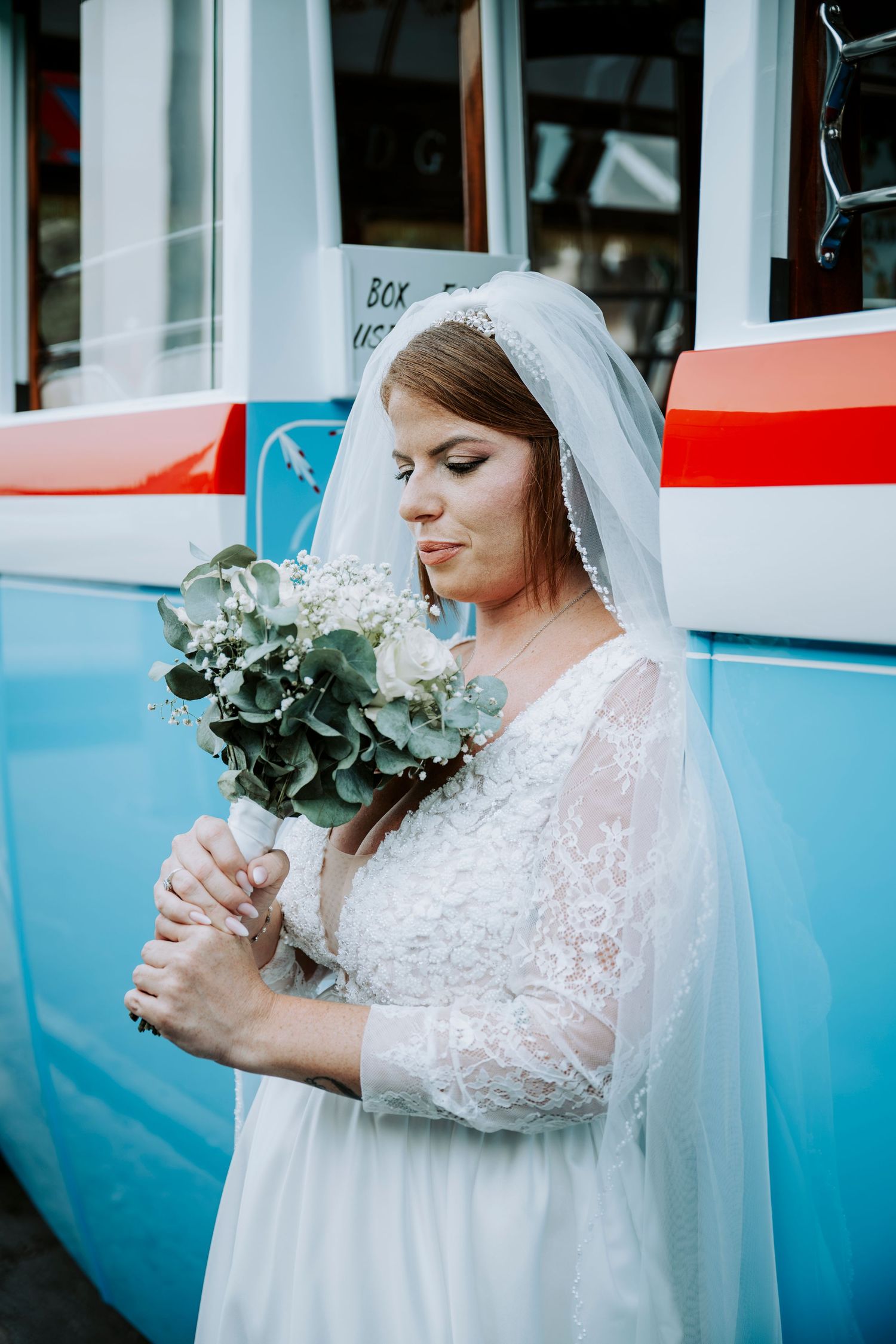 Bride in white lace dress and veil holding a green and white bouquet, standing in front of a blue vehicle.